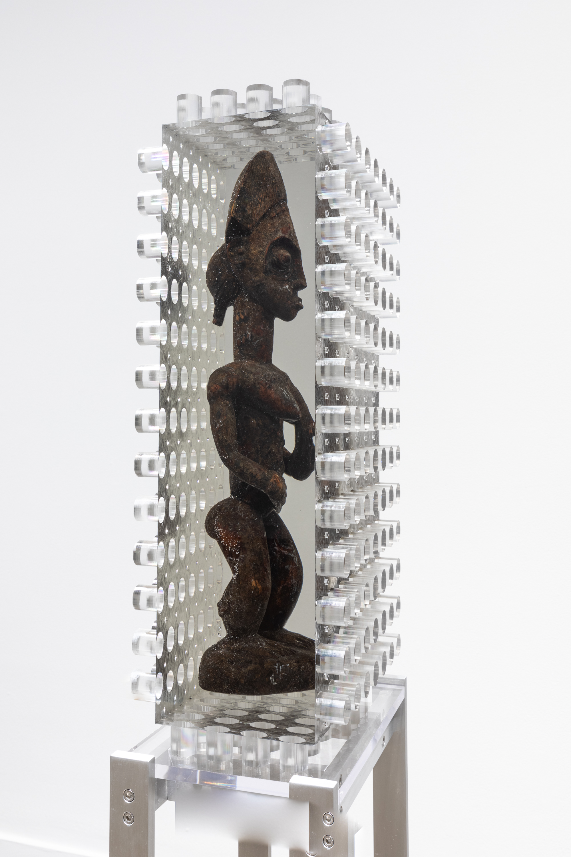 : Installation view of an African wooden sculpture of a figure encased in a block of resin with jagged edges on four of the six sides.
