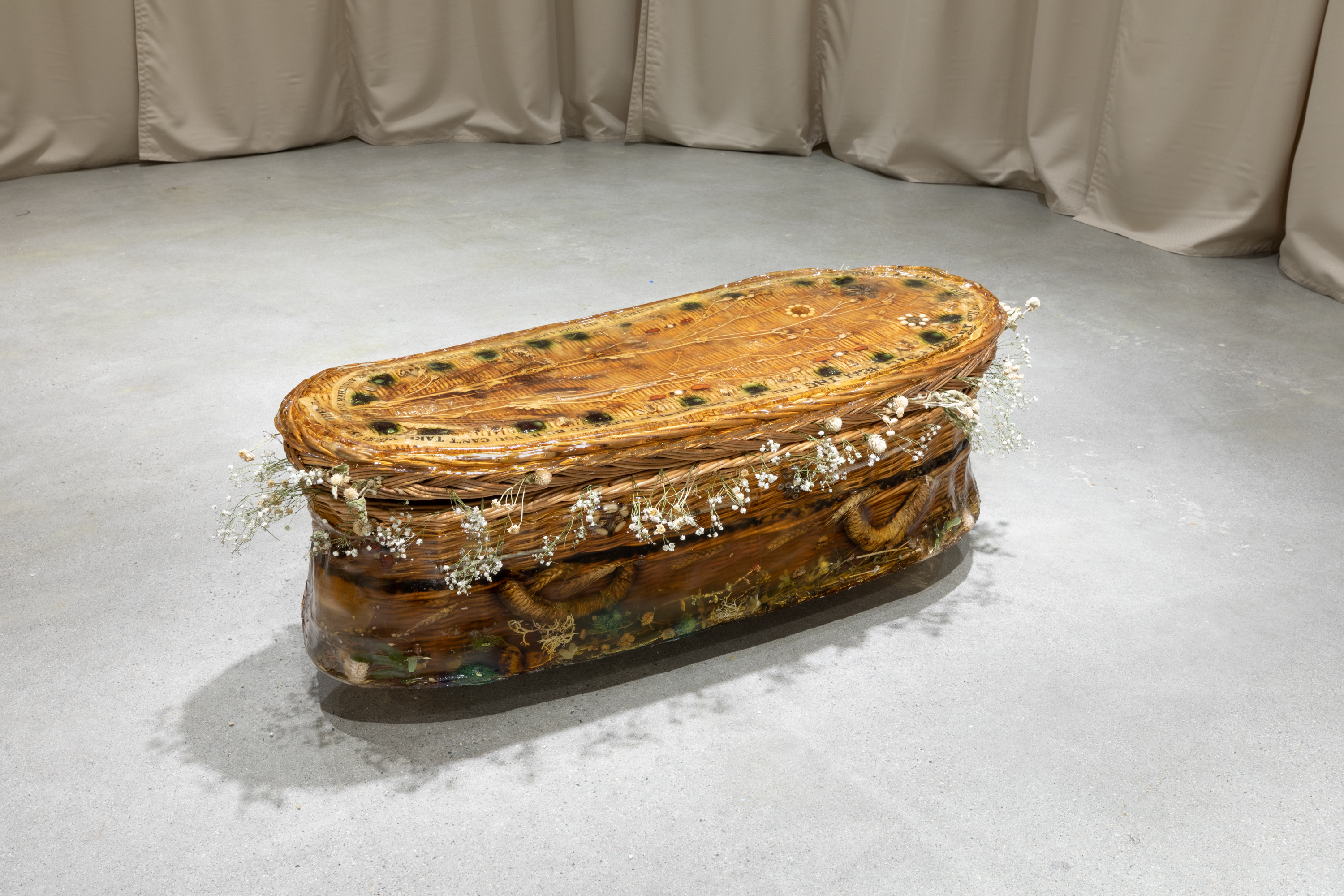 a rounded wicker casket encased in resinous gelatin sits  on the foor. Objects including syringes, pills, and beans adorn its top and lower sides, and dried flowers surround the opening between its lid and base.