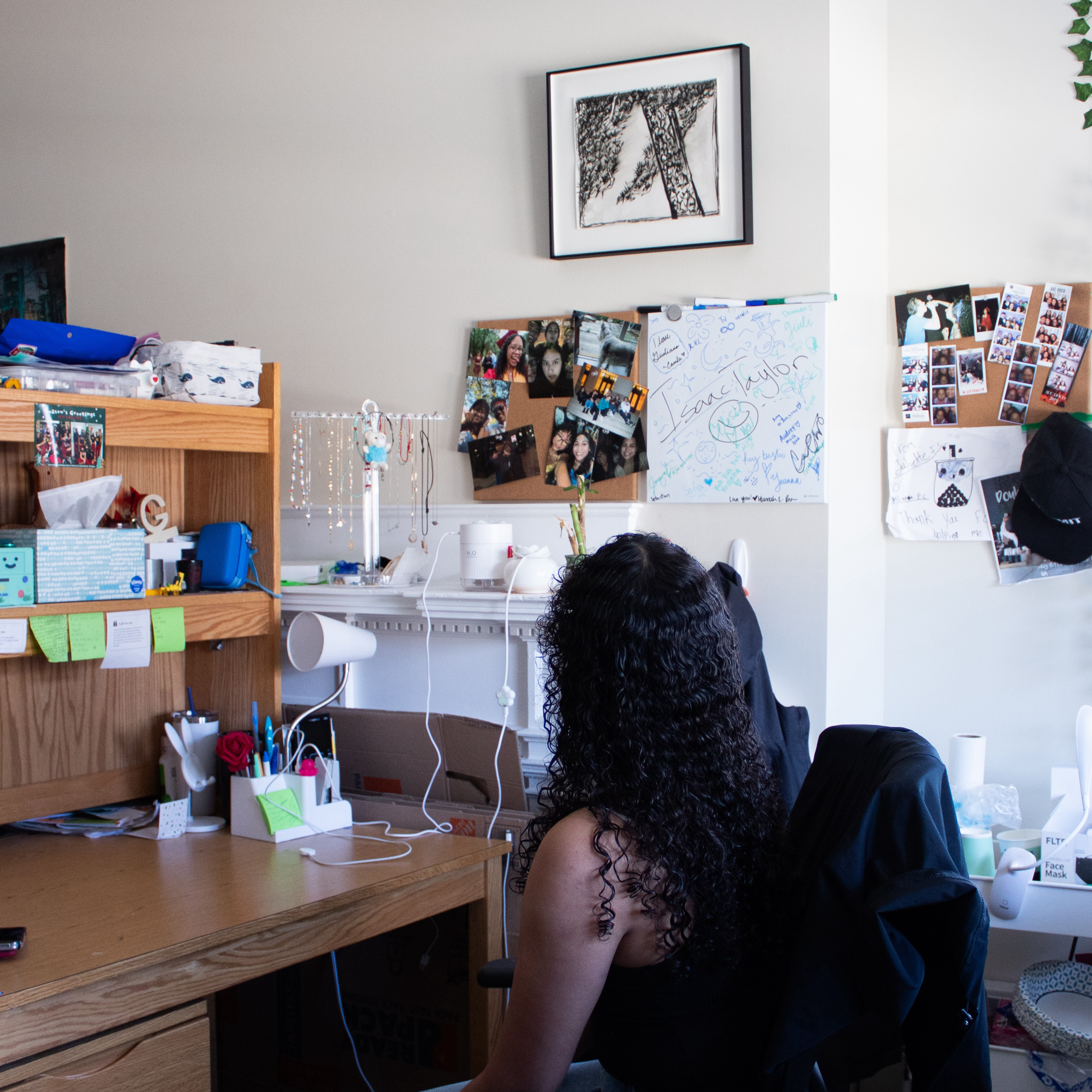 A girl sits at her desk in a dorm and looks up at a print on the wall.