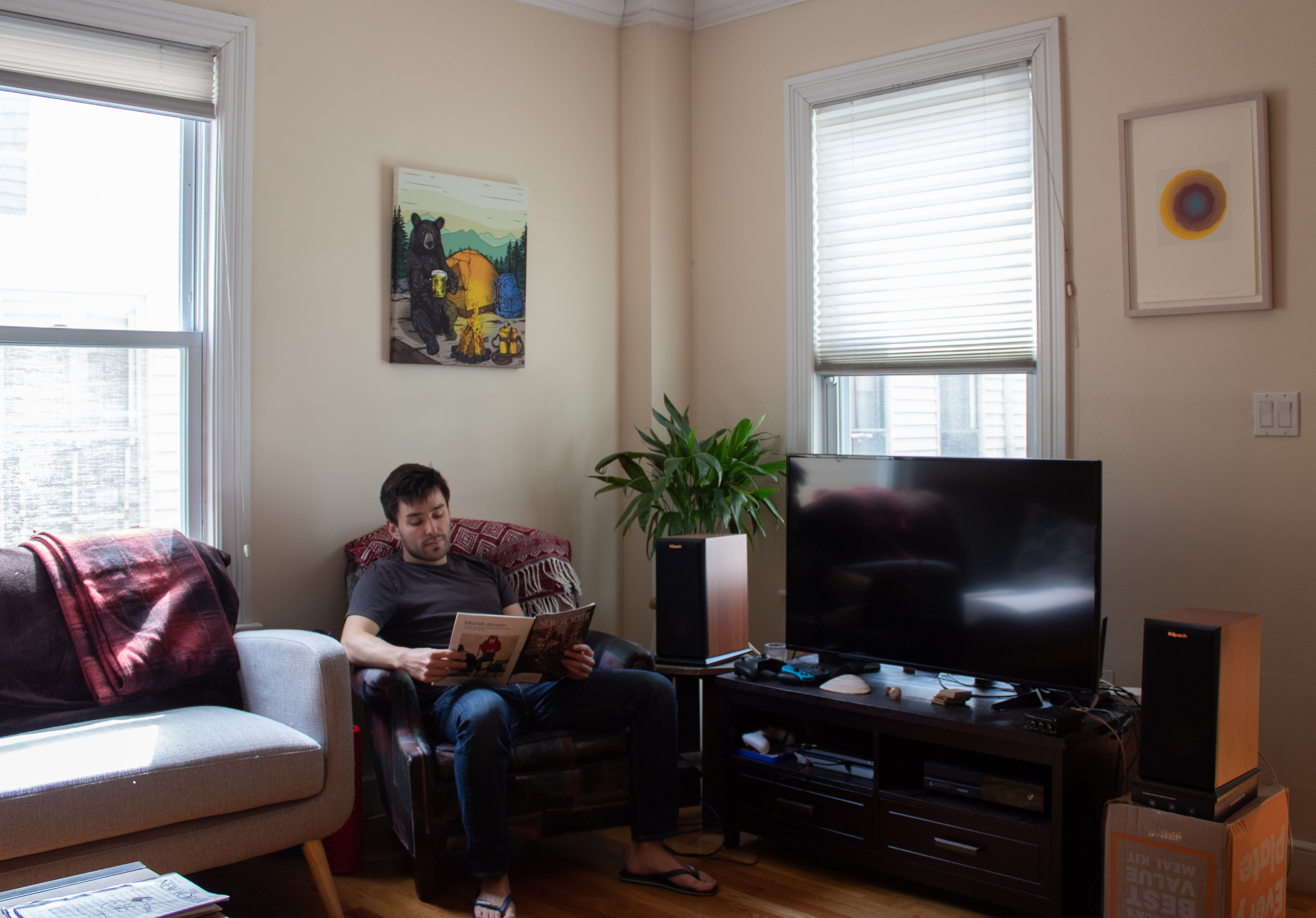 A man sits in his living room reading a magazine with two windows in the background.