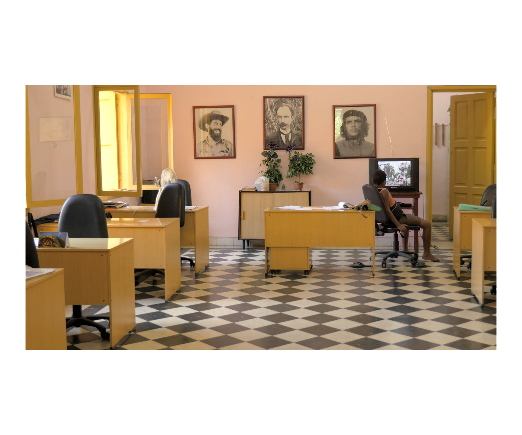 A person sits at an office desk watching television. The office has many empty desks, portraits of three men hanging on the back wall and a checkered floor.