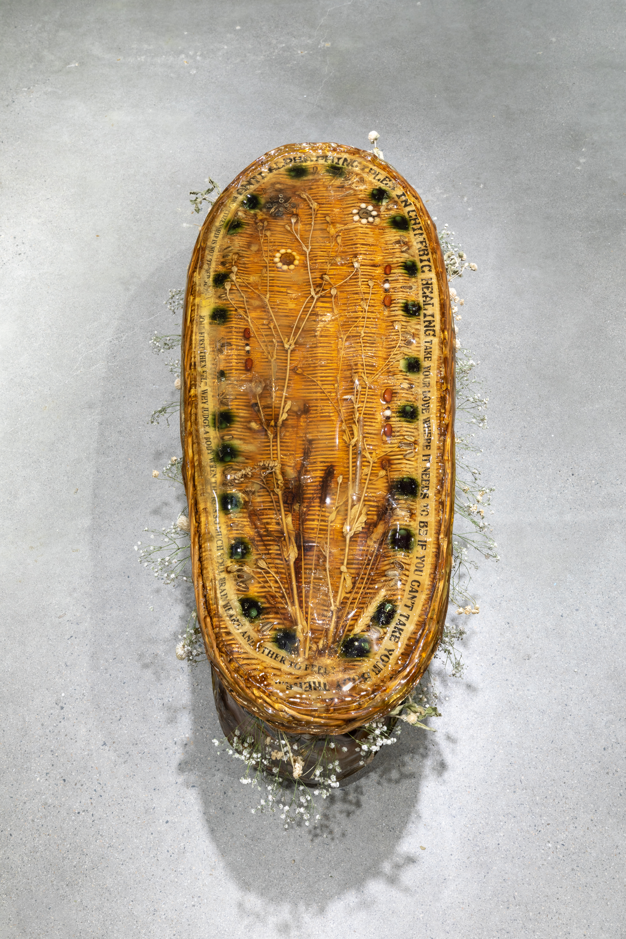 View from above of a wicker casket encased in resinous gelatin with objects including syringes, pills, and beans adorn on the top and dried flowers surround the opening between its lid and base.