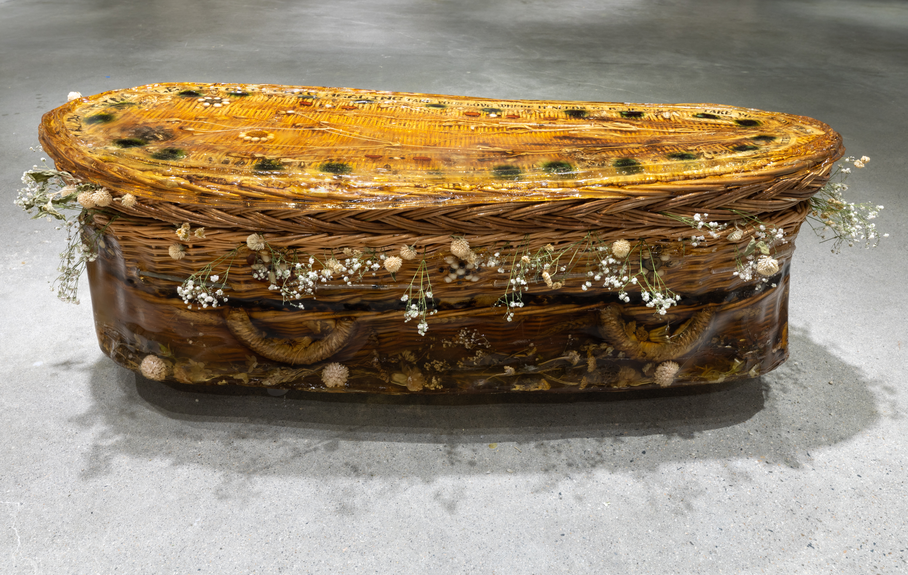 A rounded wicker casket encased in resinous gelatin sits on the floor. Objects including syringes, pills, and beans adorn its top and lower sides, and dried flowers surround the opening between its lid and base.