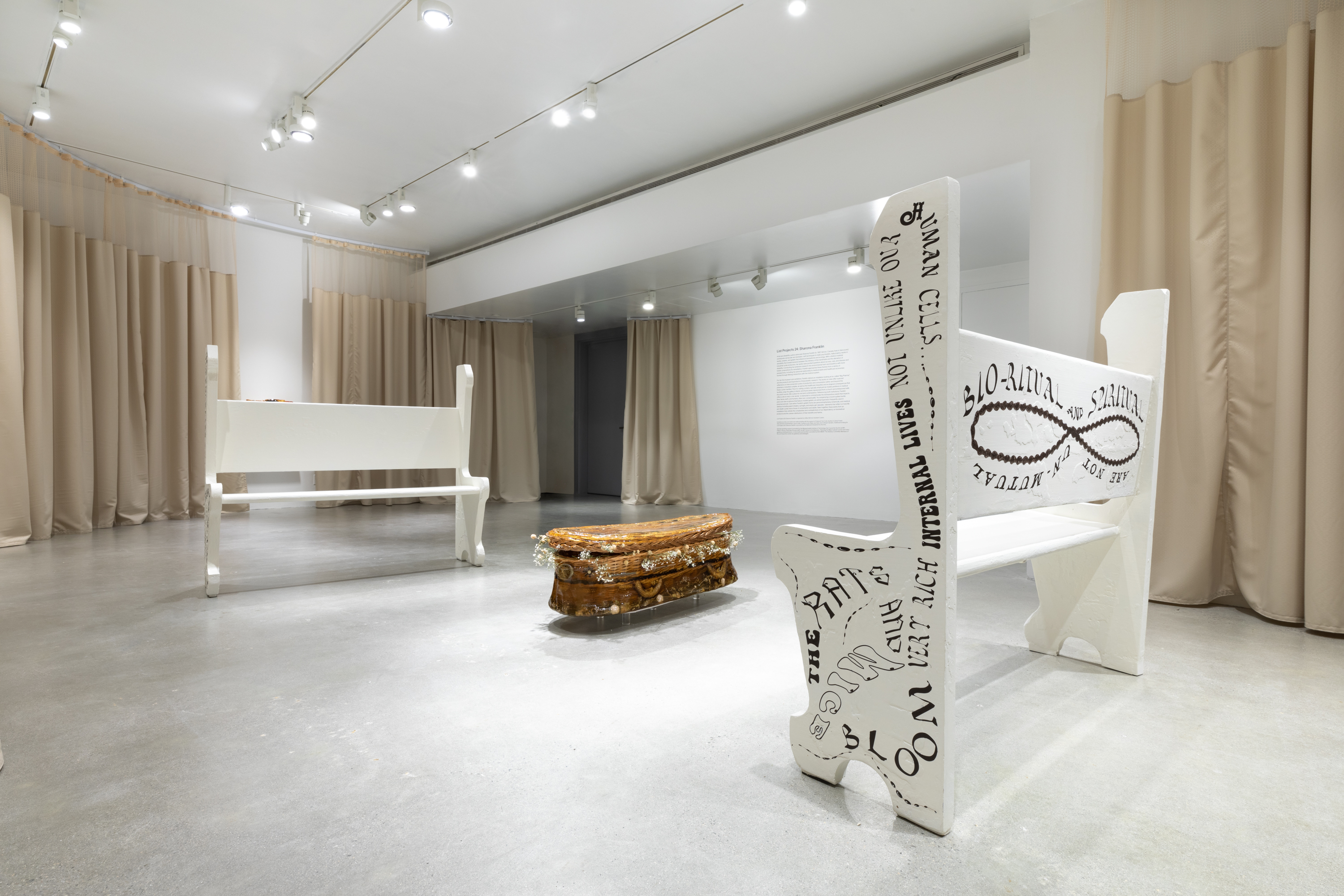 Two white church pews with text on sides and backs frame a sculpture of a wicker casket cast in gelatin with tan hospital curtains surrounding the interior of the gallery