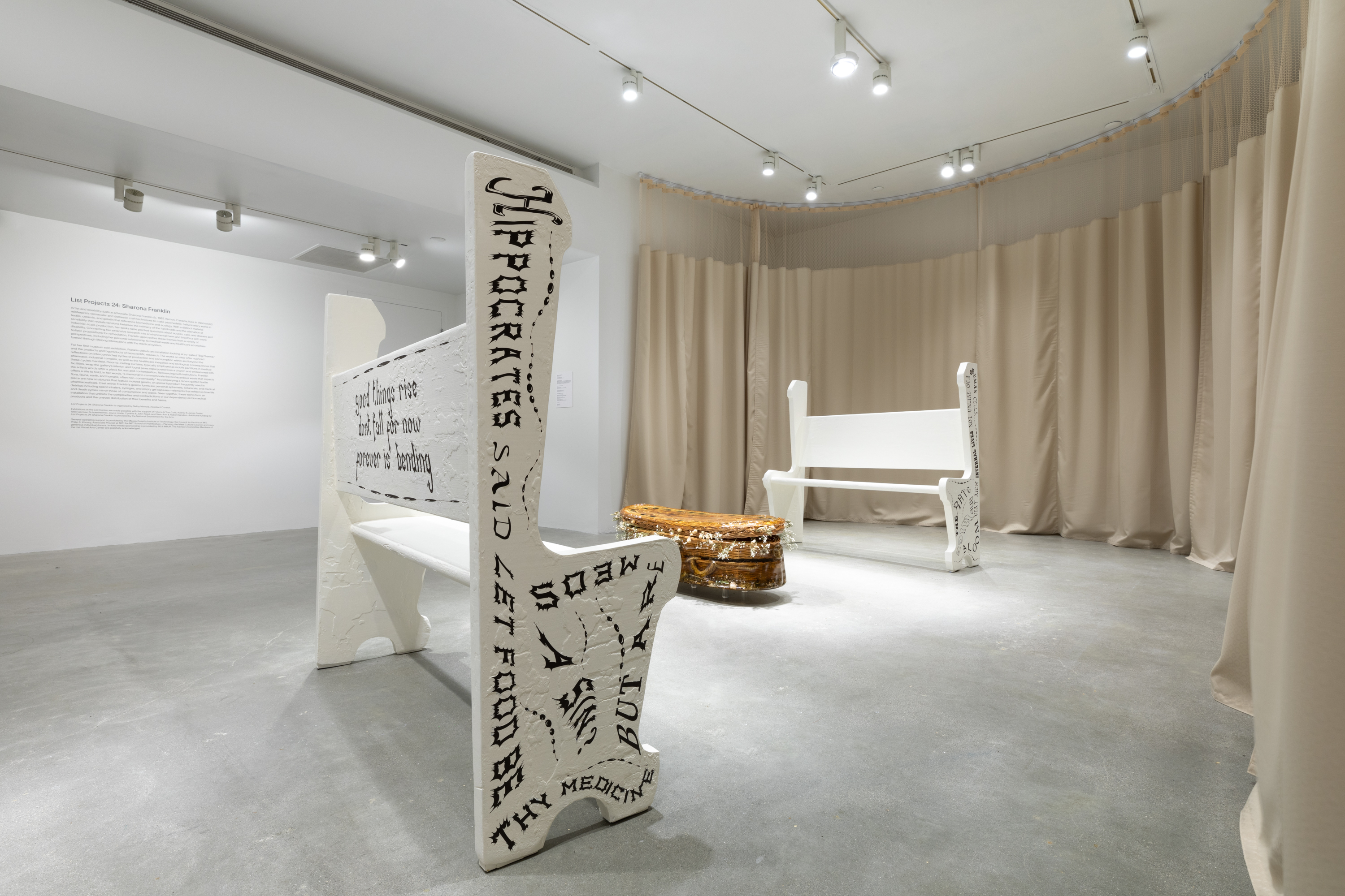 Sculpture of a willow wicker child size casket cast in gelatin sits on the floor in the middle of two white painted wooden church pews with black writing on the sides and back.