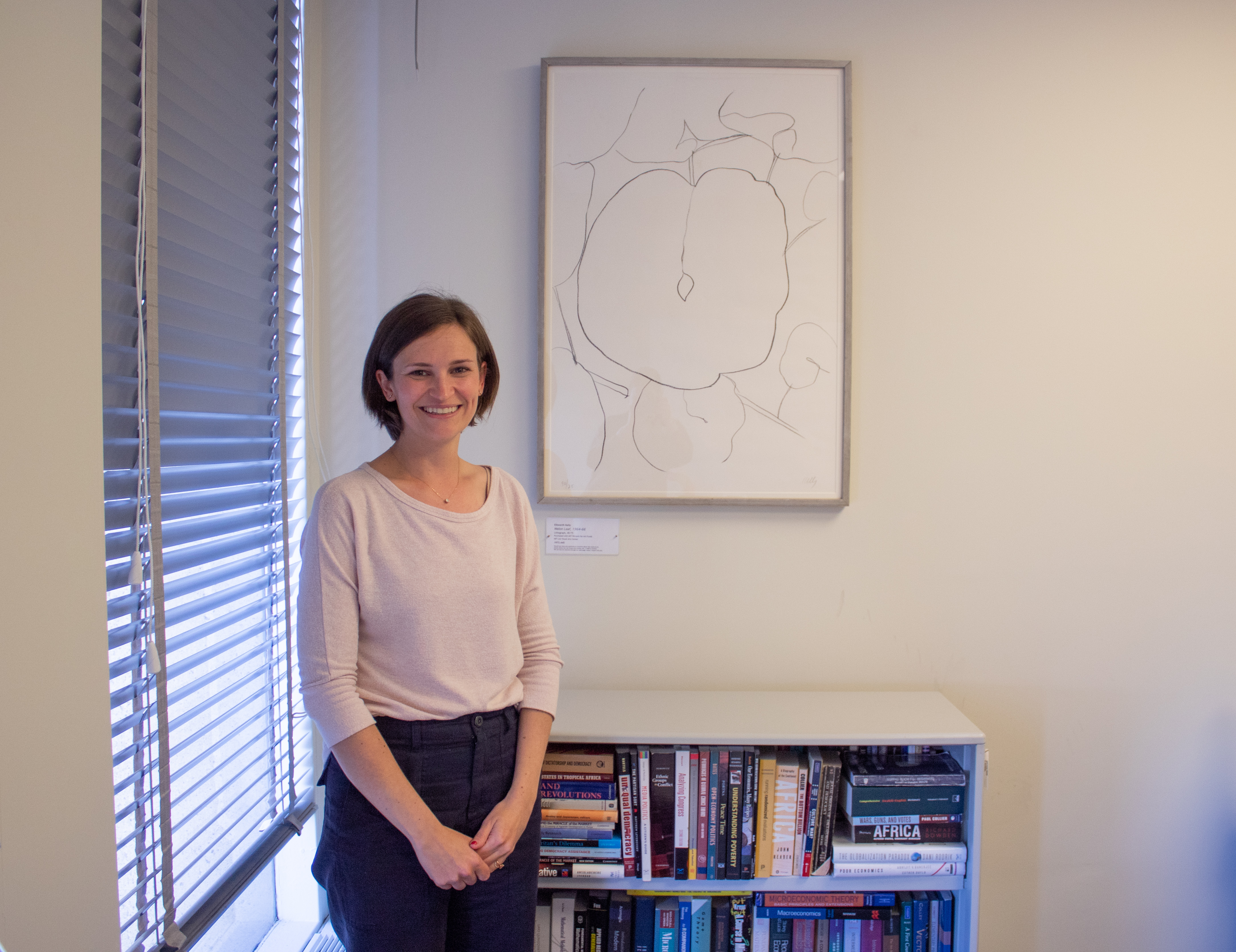 A woman in a white blouse stands in front of a bookcase with a framed print above it with her hands together smiling.