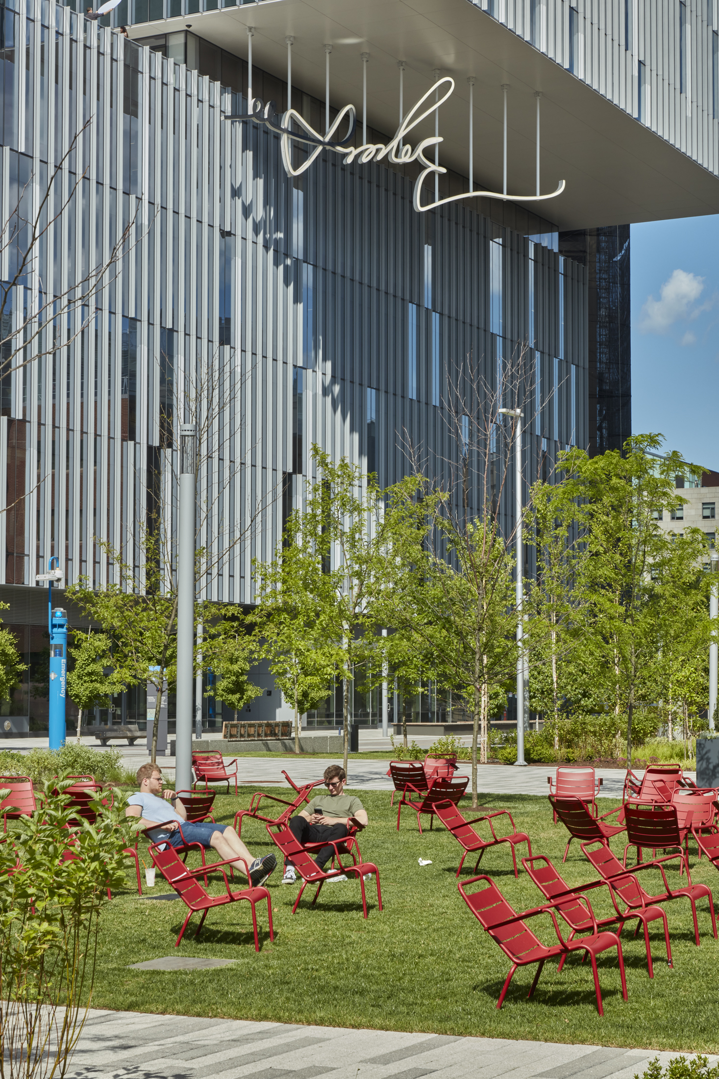 Lawn with red steel chairs in the foreground with a building in the back and newly installed LED sculpture of a hand drawn signature hanging down from the buildings overhand.
