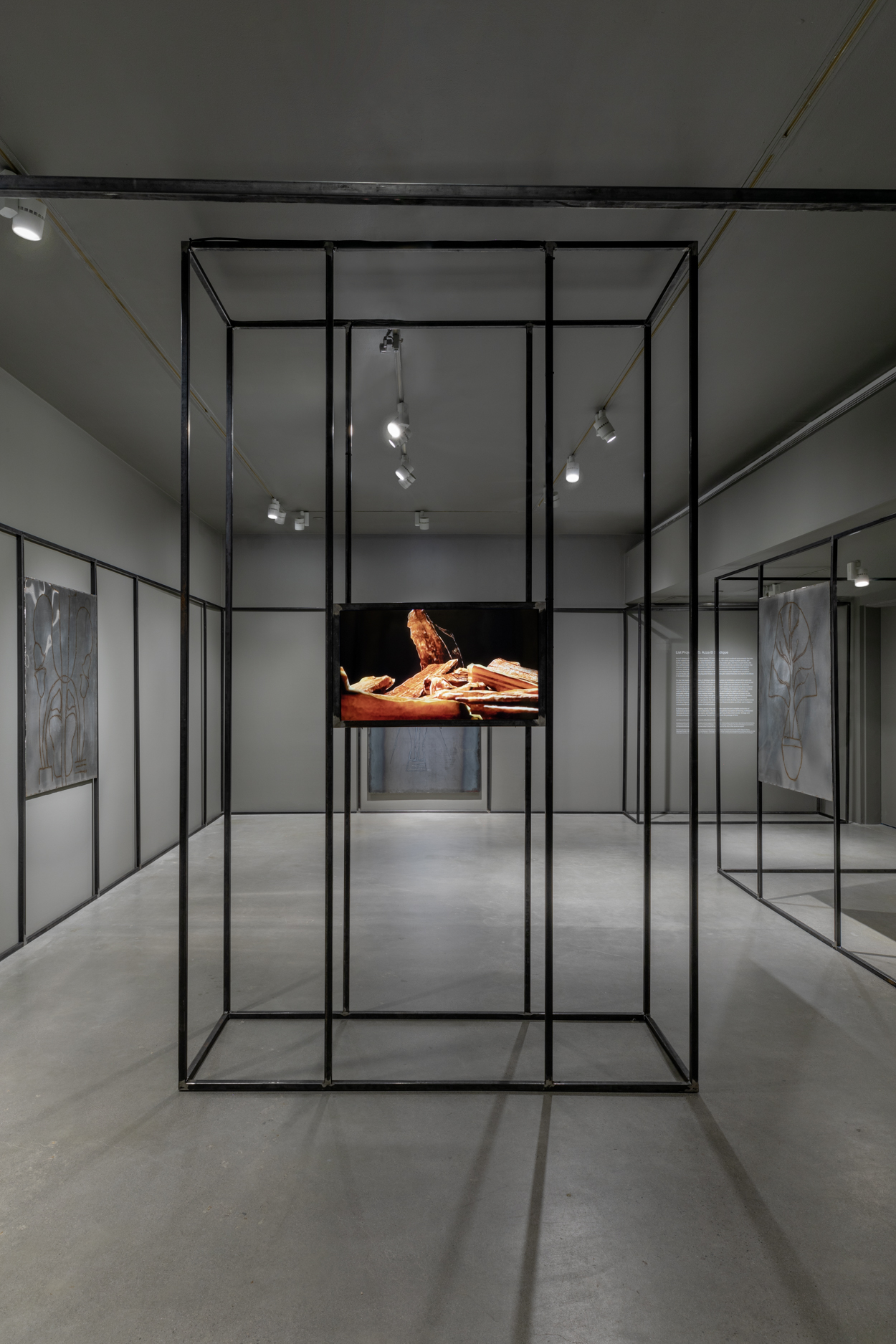 Wide angle photograph featuring a small video monitor which plays in the center of the gallery in between a large open air sculpture made of black steel bars.