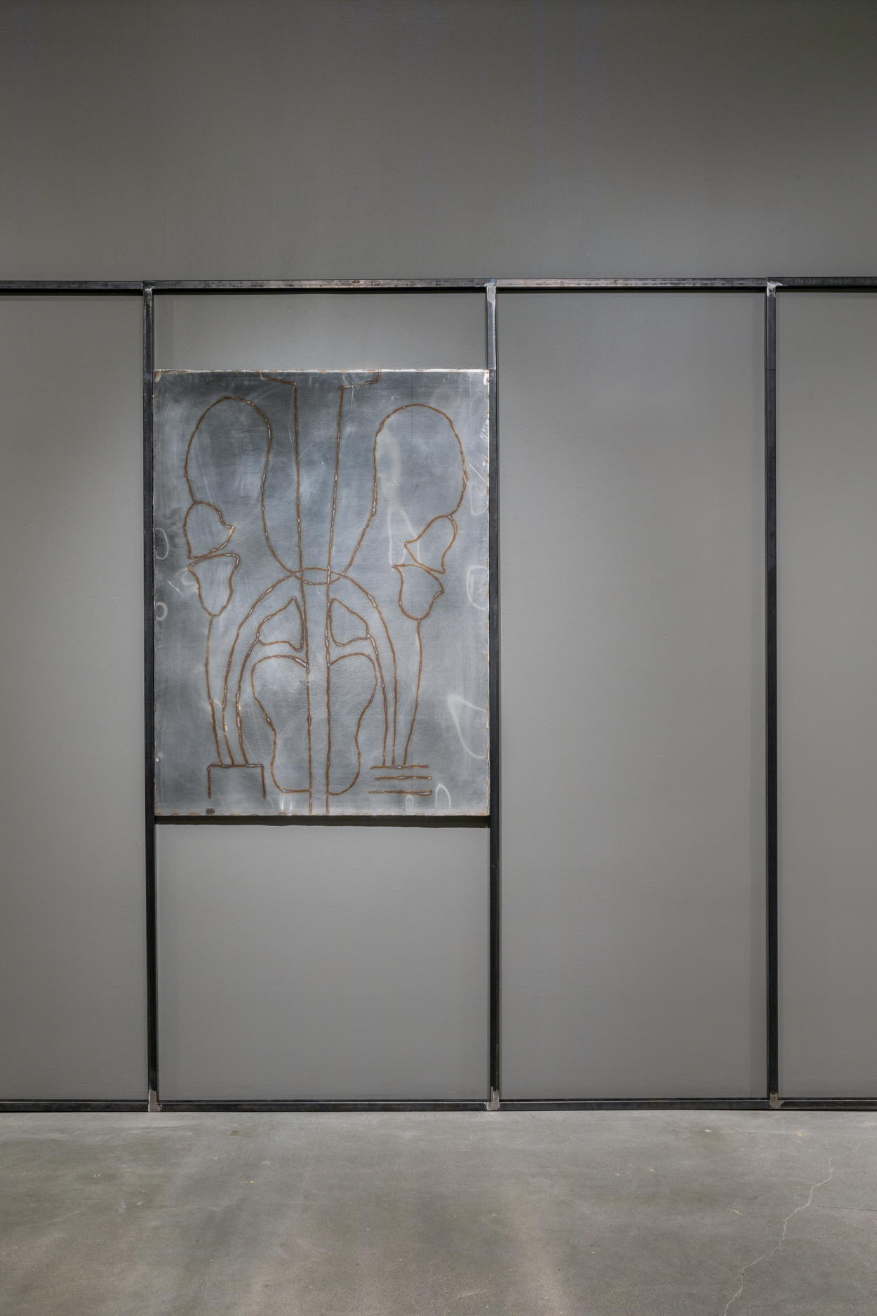 : A welded steel drawing sits in between two vertical steel bars on a gallery wall surrounded by other steel bars that form rectangular shapes on the wall.