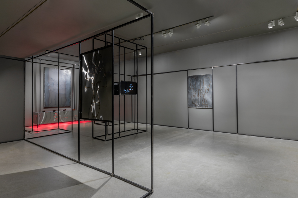 Gallery view with grey walls and cement flooring featuring a welded steel drawing on the left, right and back wall, a monitor in the center, a steel platform at rear glows red from heat lamps underneath, and black steel architecture that snakes along the periphery of the space.