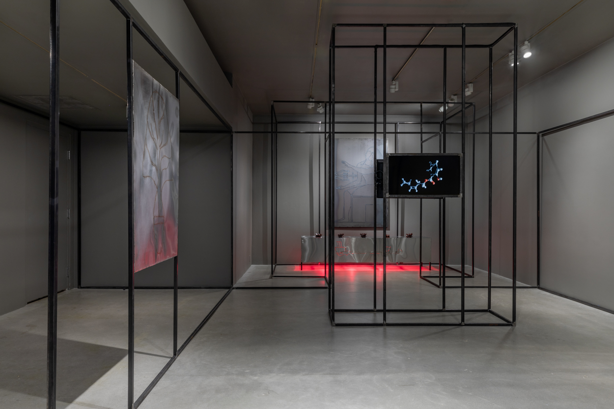 Gallery view with grey walls and cement flooring featuring welded steel drawings on the left and right, a monitor in the center, a steel platform at rear glows red from heat lamps underneath, and a black steel architecture snakes along the periphery of the space.