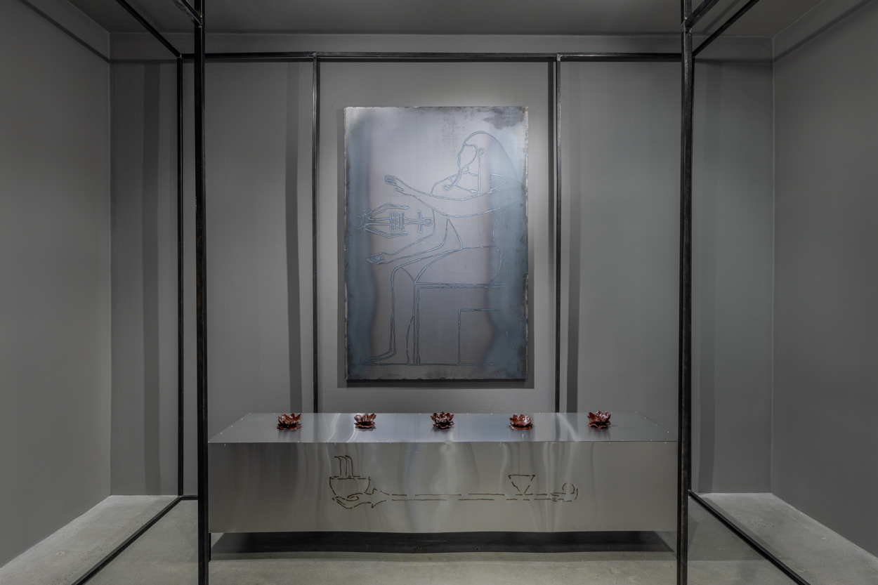 Black steel architecture frames a metal table against the rear gallery wall with five small bukhoor sculptures on top with a large, welded steel drawing of a Pharaoh hanging above the table.
