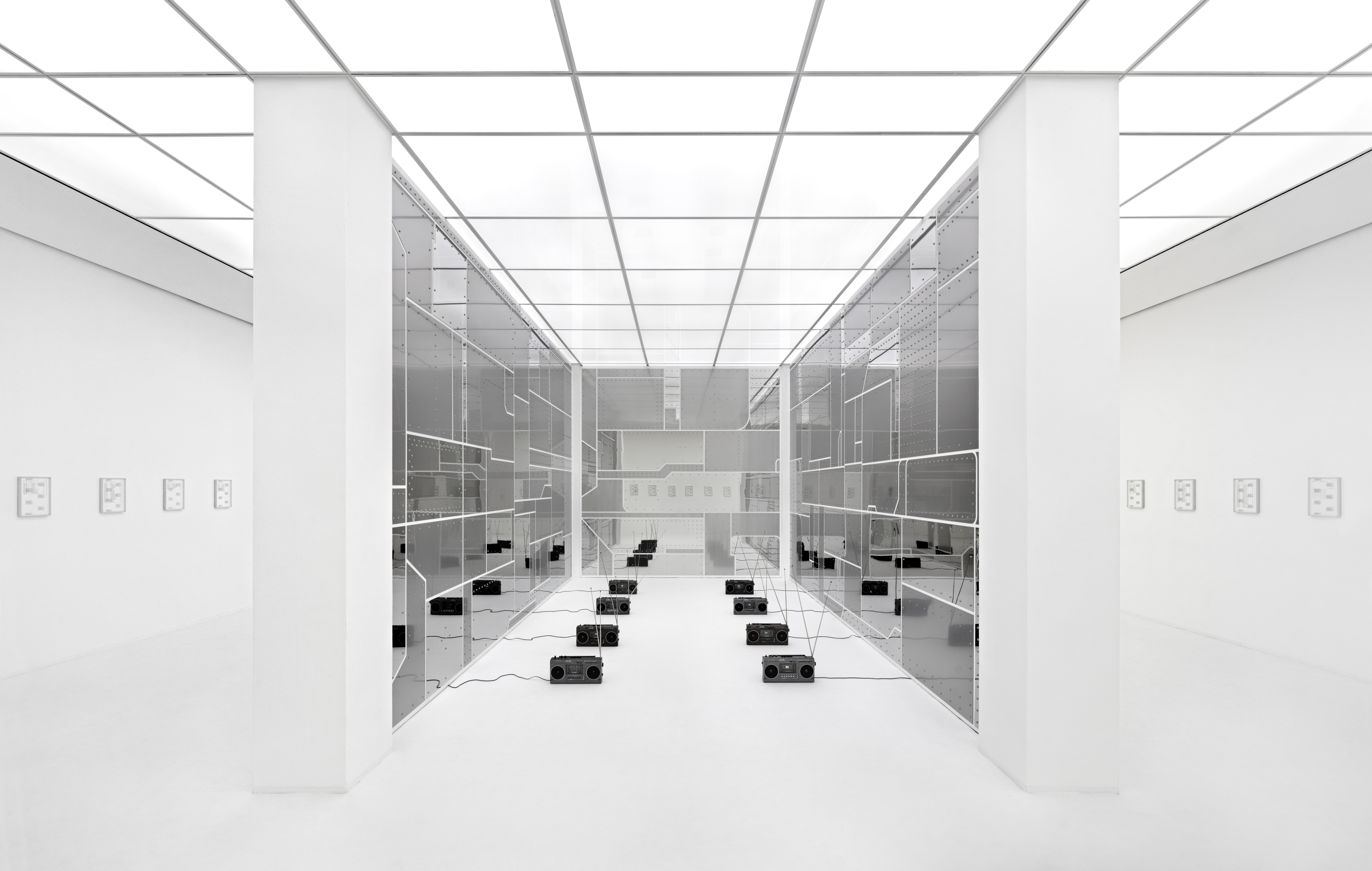 Symmetrical installation view of a black and white gallery with small prints hung on the left and right walls and an installation in the center of the room with mirrors cut out to resemble airplane parts frames GDR-era radios placed on the floor