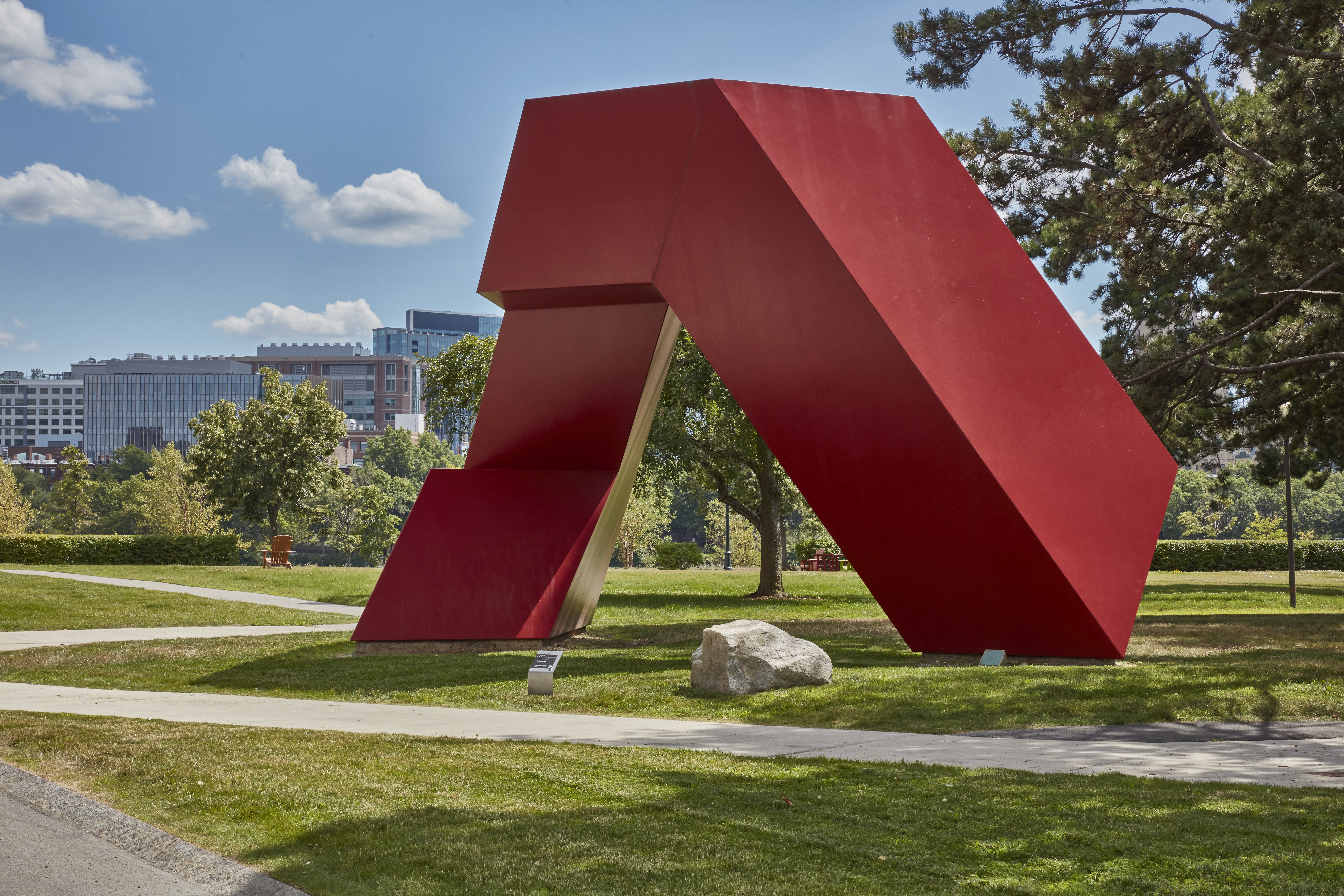 Red painted steel geometric sculpture with hard edges and rectangular shapes within on a grass field.