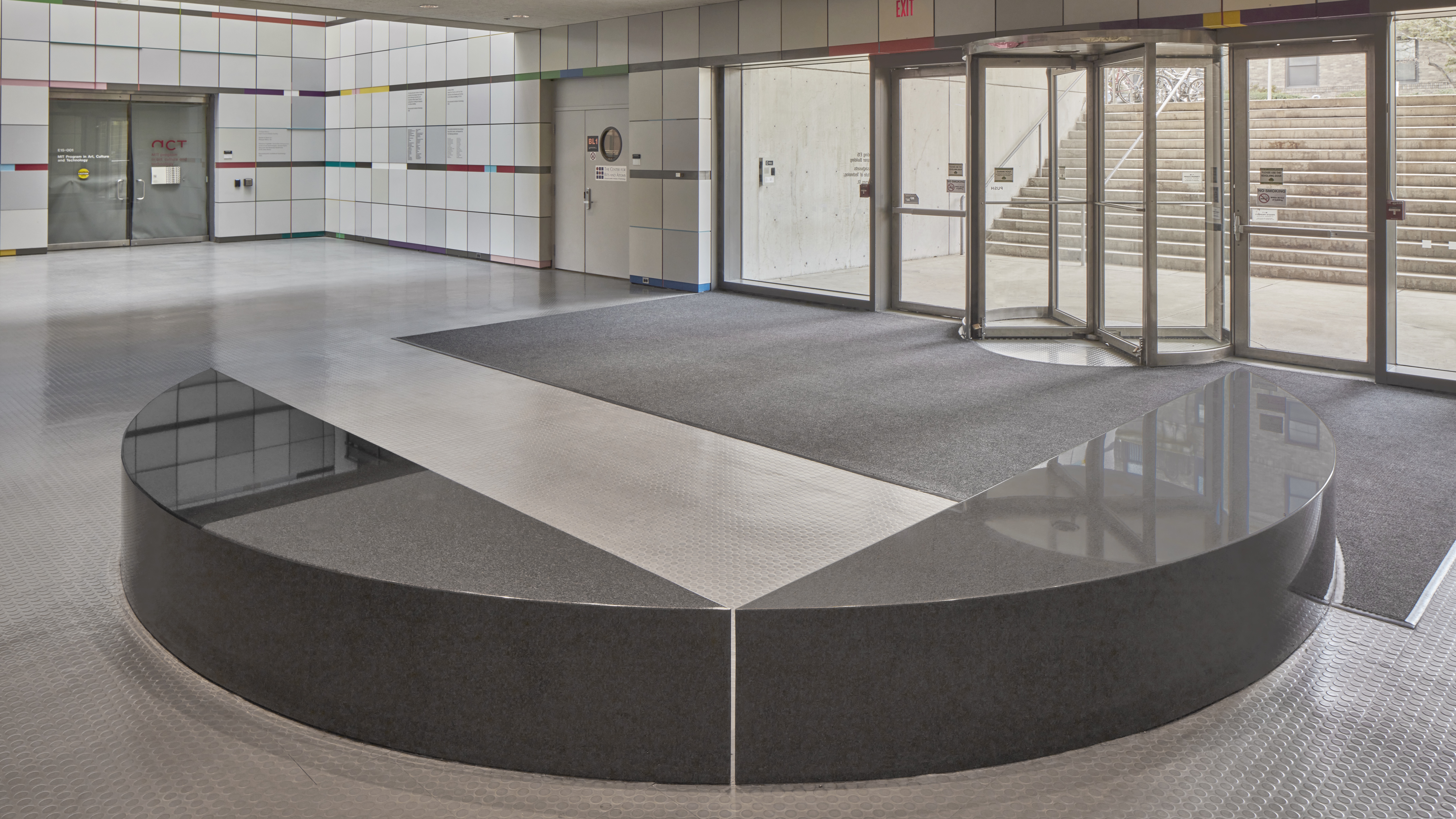 L-Shaped granite bench with two rounded edges facing a set of revolving doors to the entrance to the building.