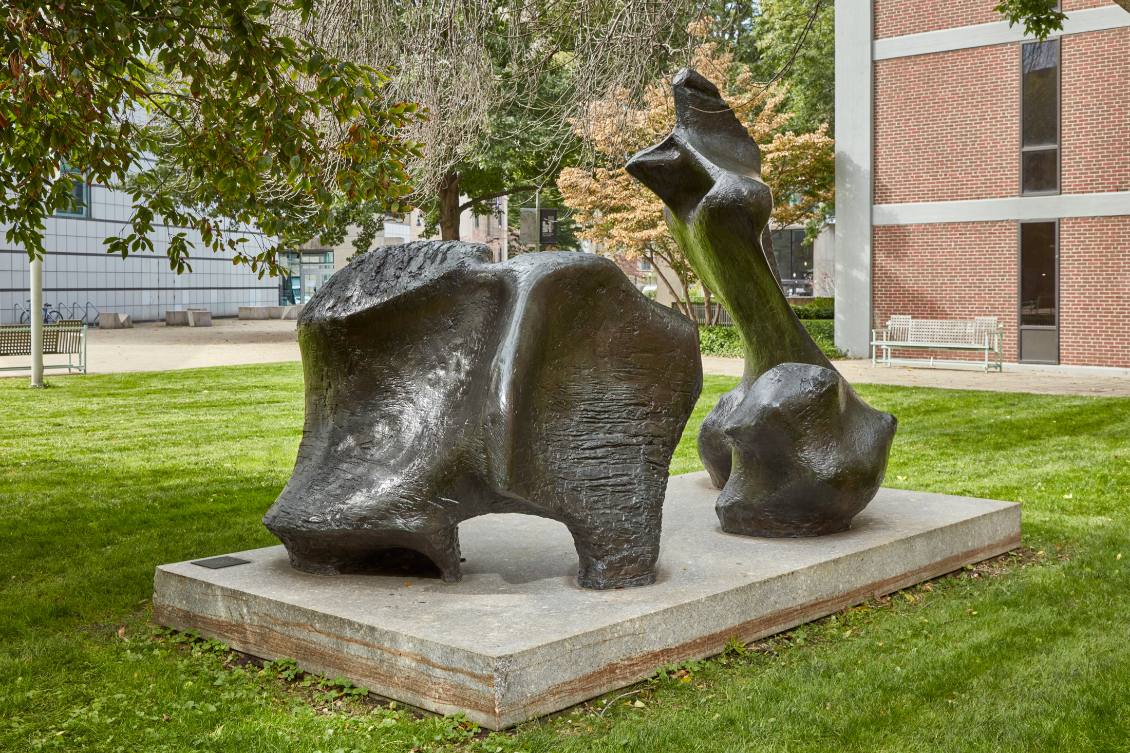 Large-scale bronze abstract sculpture of a figure on a stone pedestal in a grassy courtyard.