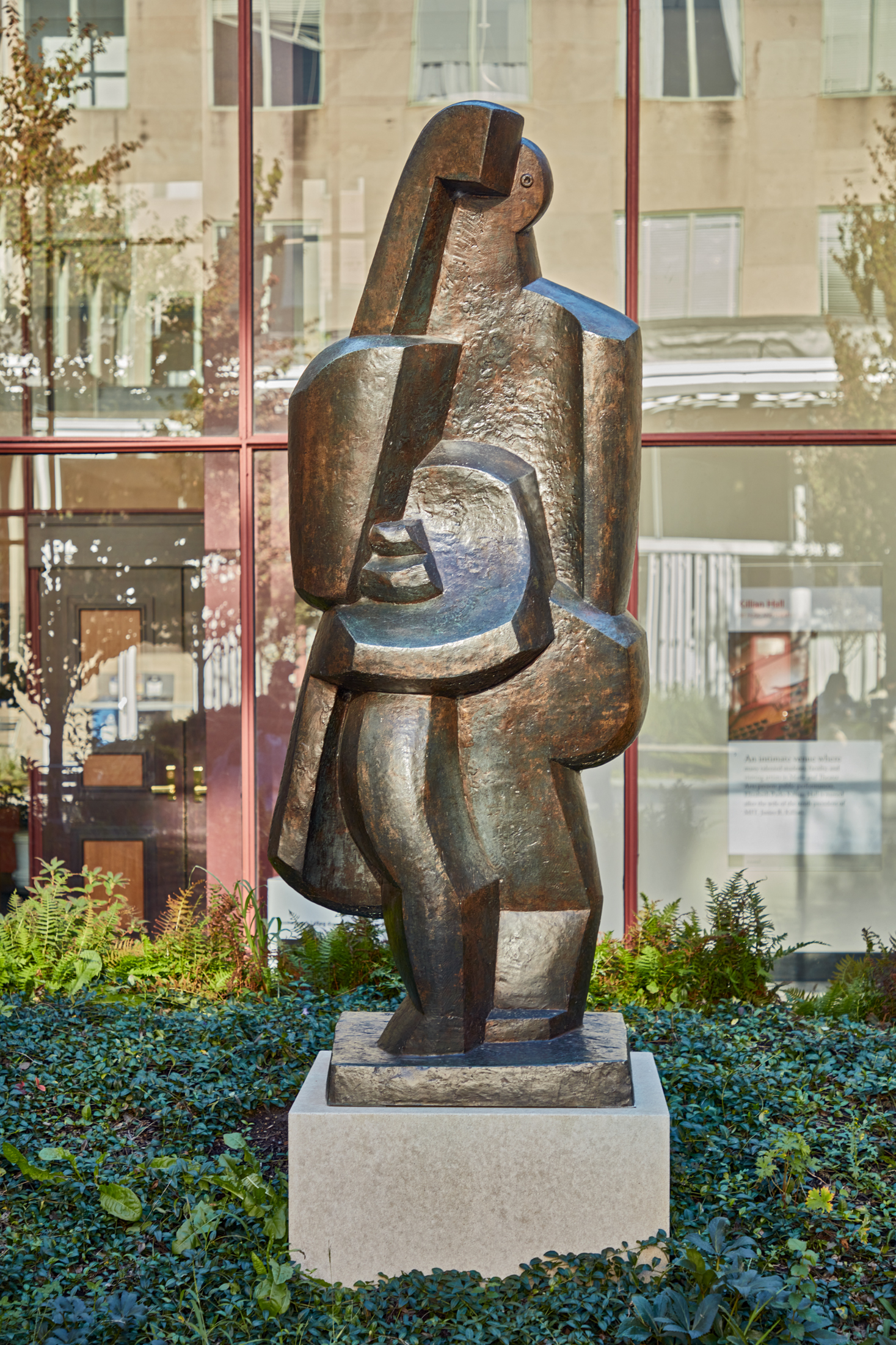 Side view of a bronze sculpture of a figure on a stone pedestal in a grassy courtyard.
