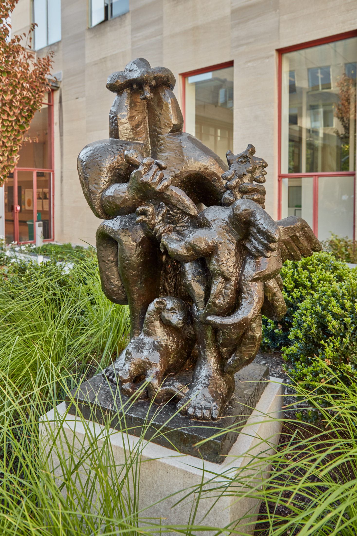 Bronze sculpture of figured tangled between one another on a stone pedestal in a grassy courtyard.