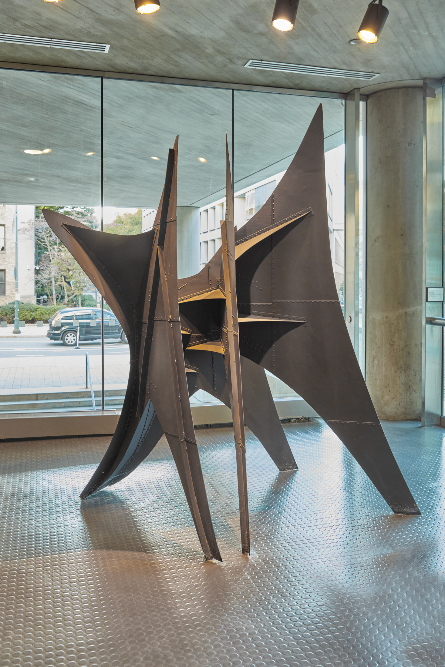Downsized model of a black steel sculpture with four points of contact on the ground and arched edges that resemble the shape of a sail.