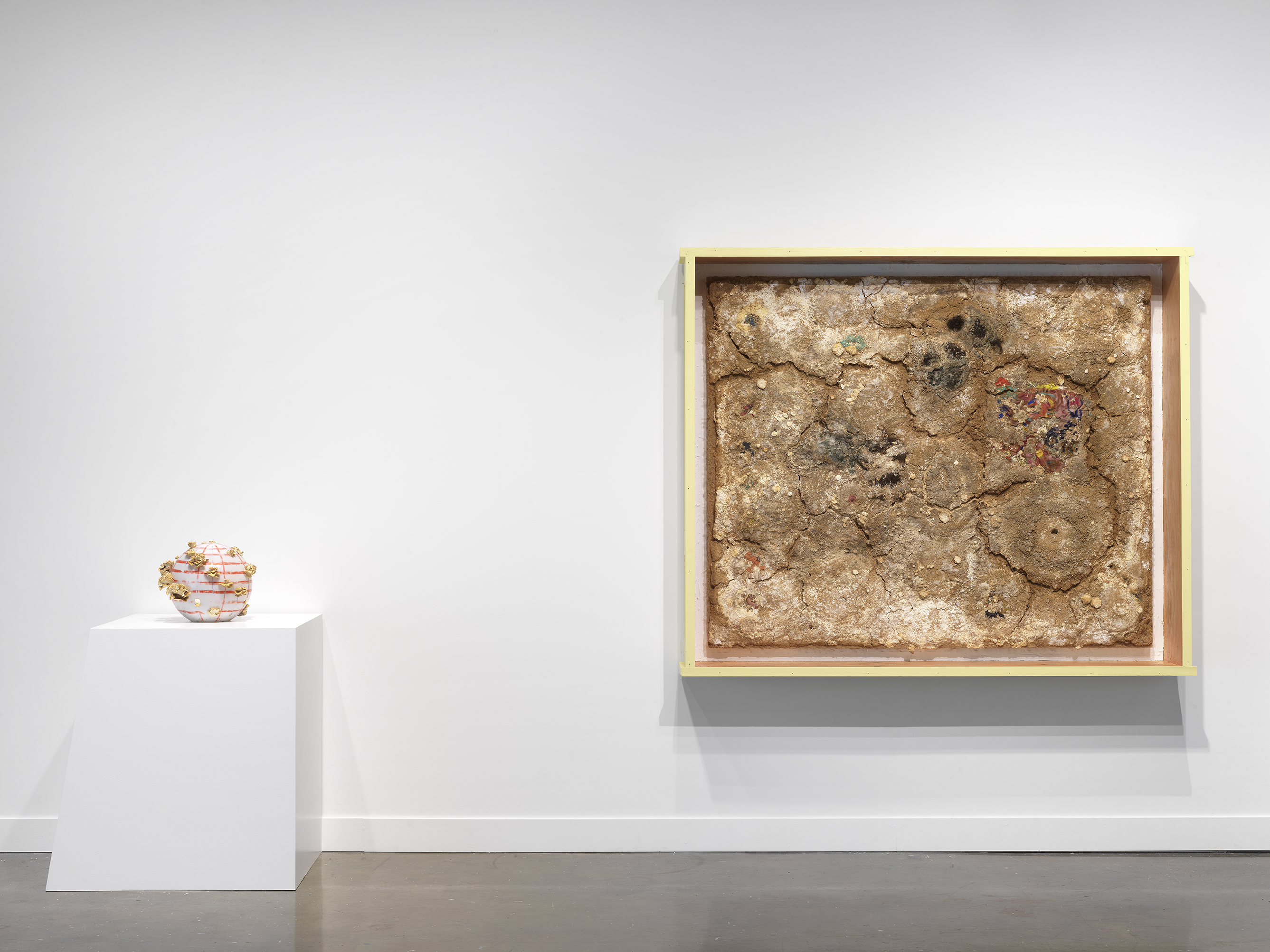 An orange and white striped beach ball with mushrooms growing on the sides sits on a geometric white pedestal sits to the left of a crusty framed artwork made of various materials resembling dried dirt.