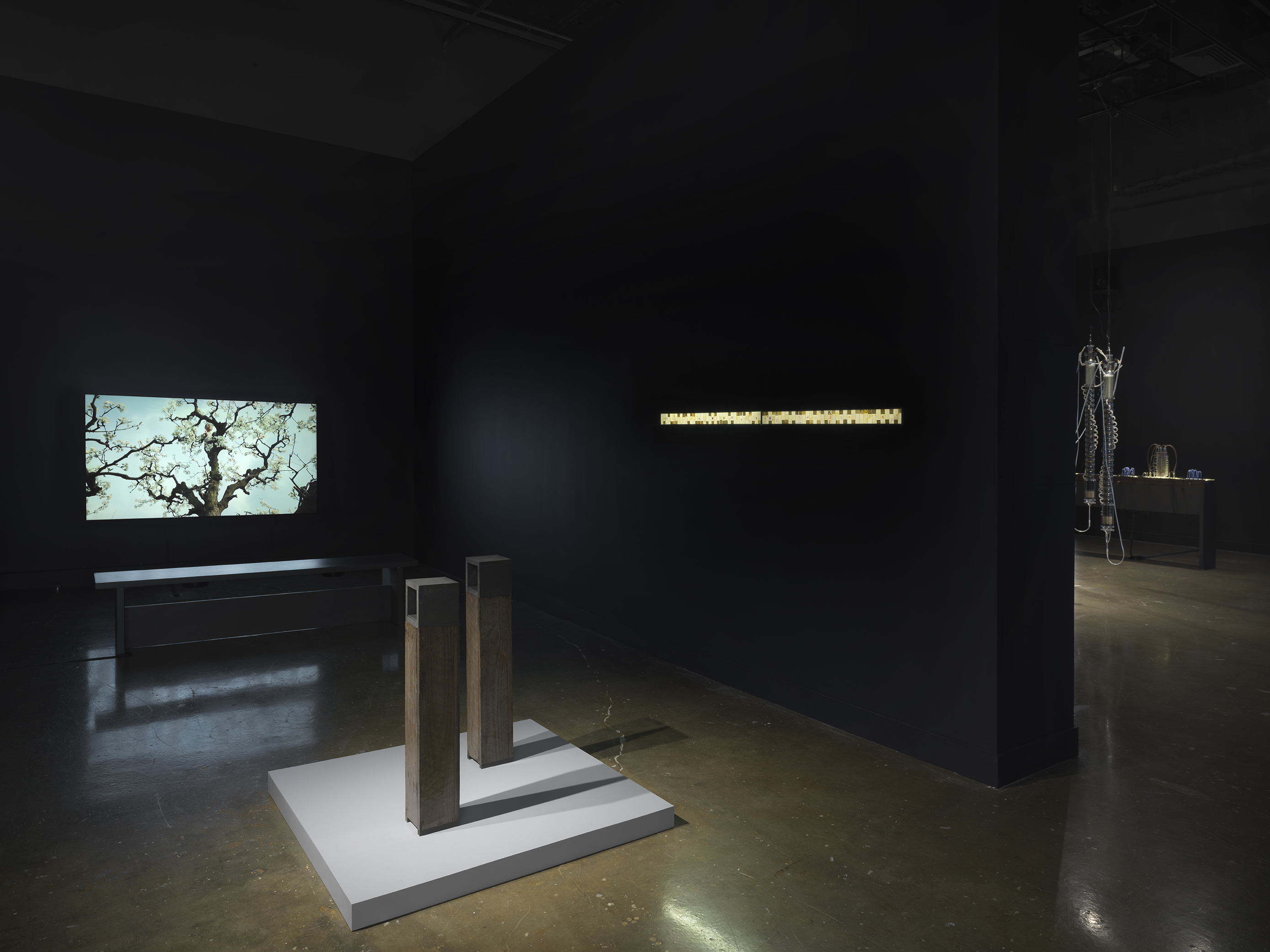 : Two slim brown pedestal sculptures surrounded by a single-channel video and a backlit yellow installation inlaid into the wall.