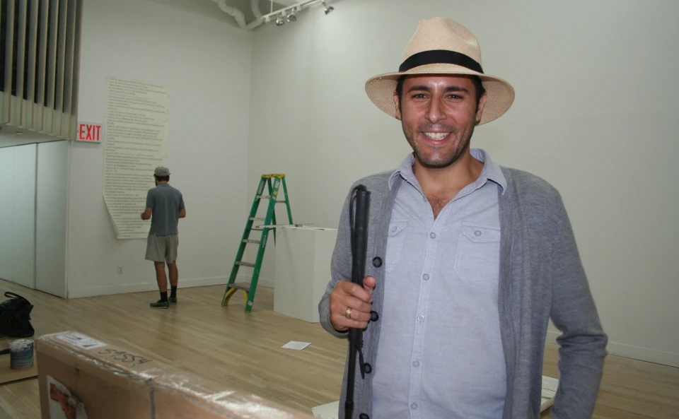 A person wearing a light blue buttoned up shirt, an open gray cardigan and a cream hat, smiles at the camera. To the right, behind them another person faces the wall next to a green latter