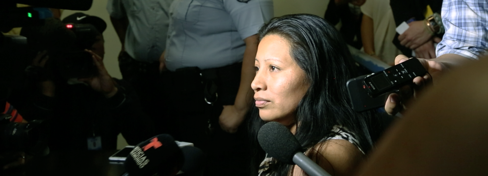 Video still of a woman with dark hair surrounded by a camera crew and microphones with her gaze to the left.