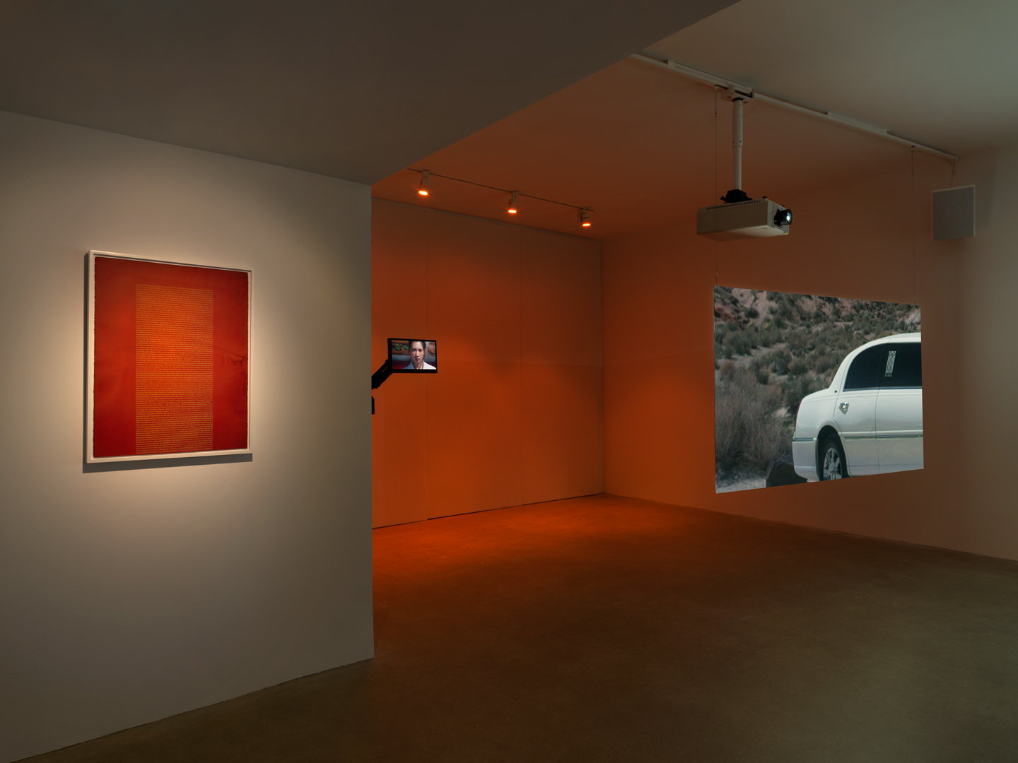 A burnt orange screen print is hung on the left wall. A screen held by a mount that looks like a car’s sideview mirror showing woman with black hair gazing at the viewer is hung off the wall to the right, lit with orange gallery lighting. To the far right, a projector shows the rear half of a white limousine against a desert landscape.