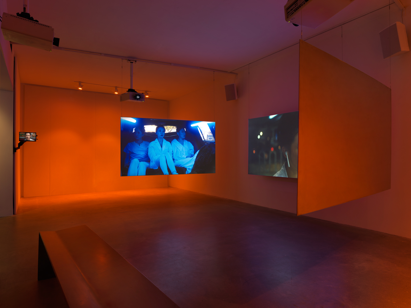Two adjacent screens show three women dressed in white sitting in the backseat of a car while the other two screens are only visible from the back.