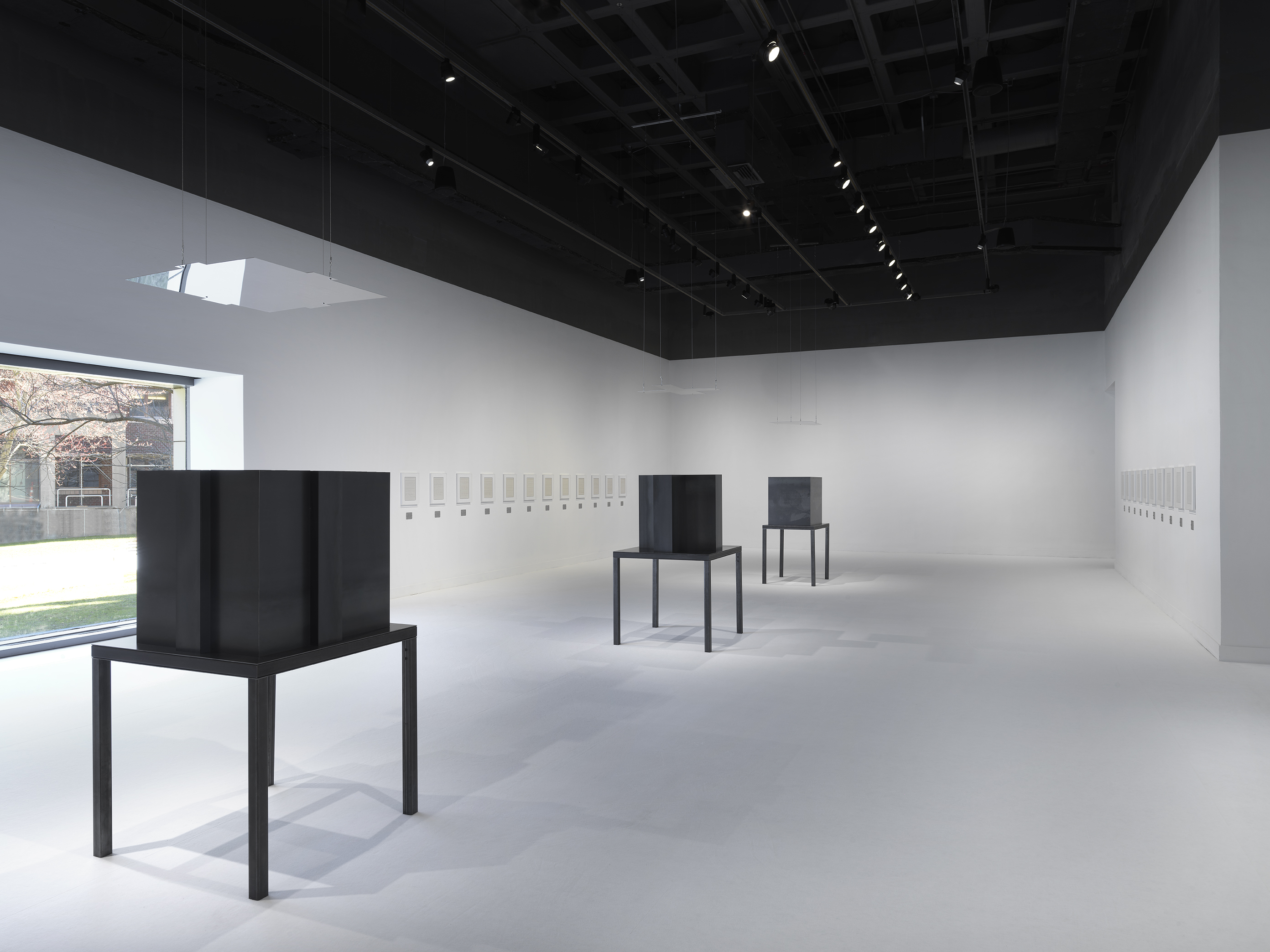 Three dark steel tables and geometric sculptures are surrounded by artworks in white frames hung horizontally in a white walled and floored gallery.