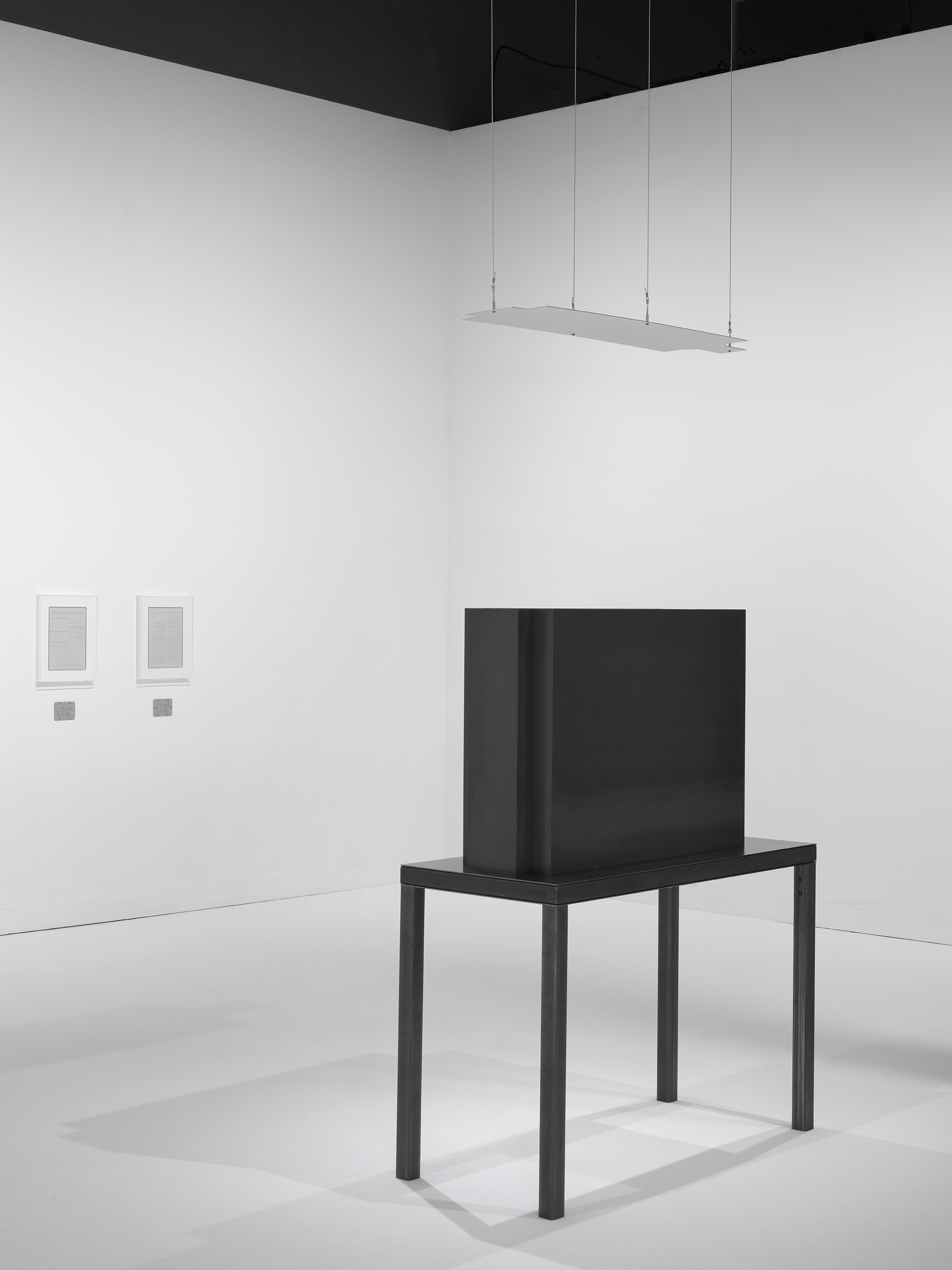 A dark steel table and geometric sculpture with a suspended mirror is opposite two framed works in a white walled and floored gallery.