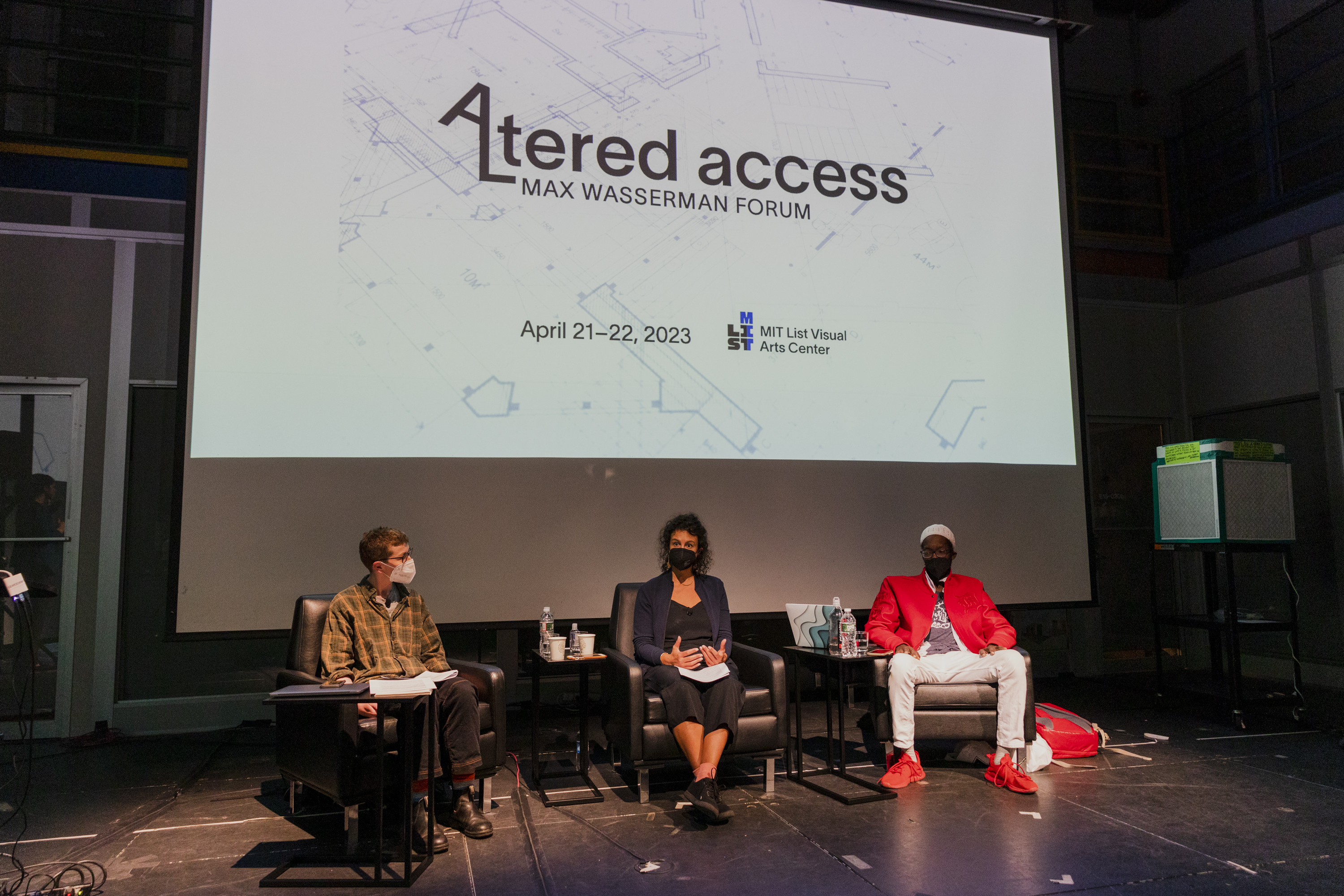 Three Altered Access participants sit on a stage in a panel discussion. Behind them is a bright projector screen that reads &quot;Altered Access, 2023 Max Wasserman Forum&quot; with the forum dates below and the MIT List Visual Arts Center logo.