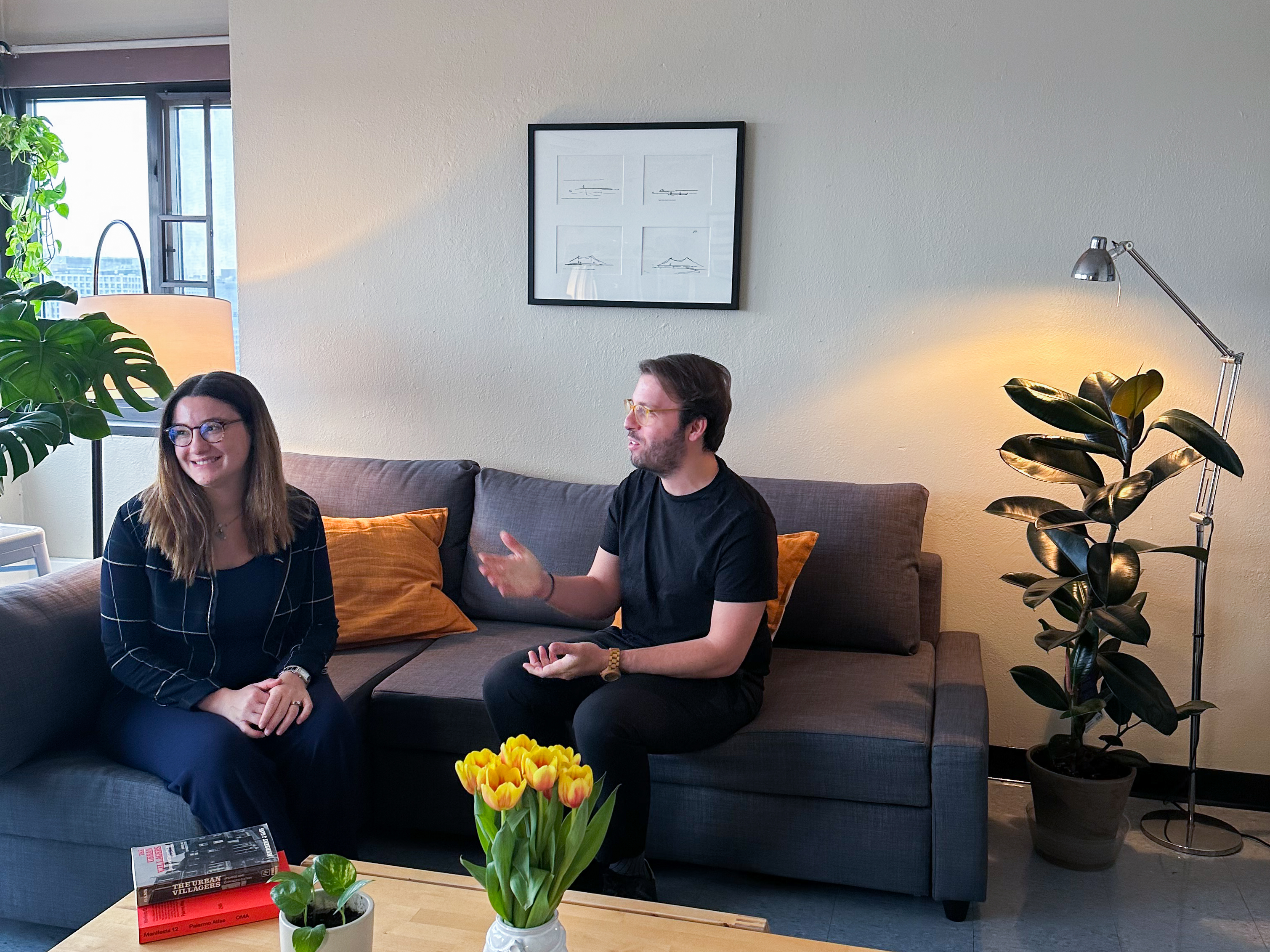 A man and woman sitting on a gray couch with a framed artwork on the wall behind.