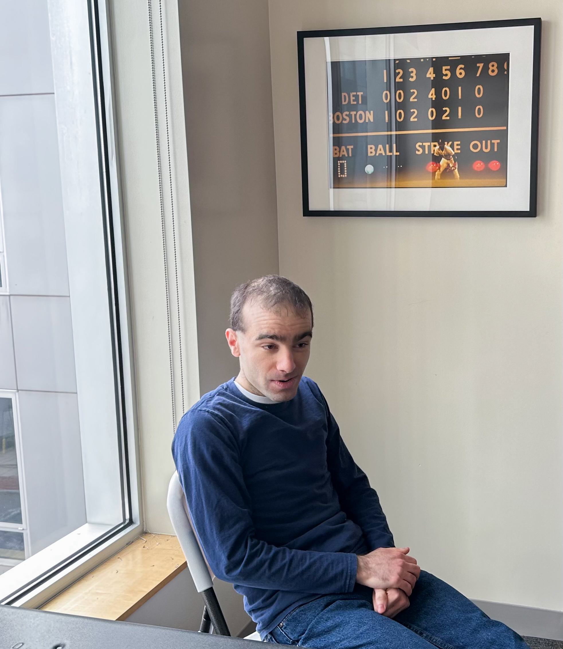 Man wearing a blue sweater sitting in chair to the left of a window. There is a black framed photograph hanging on the wall behind the man.