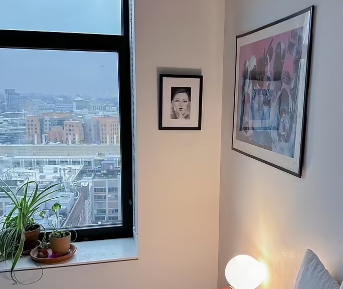 Two framed works of art hanging on a wall to the right of a large window with several plants on the windowsill.