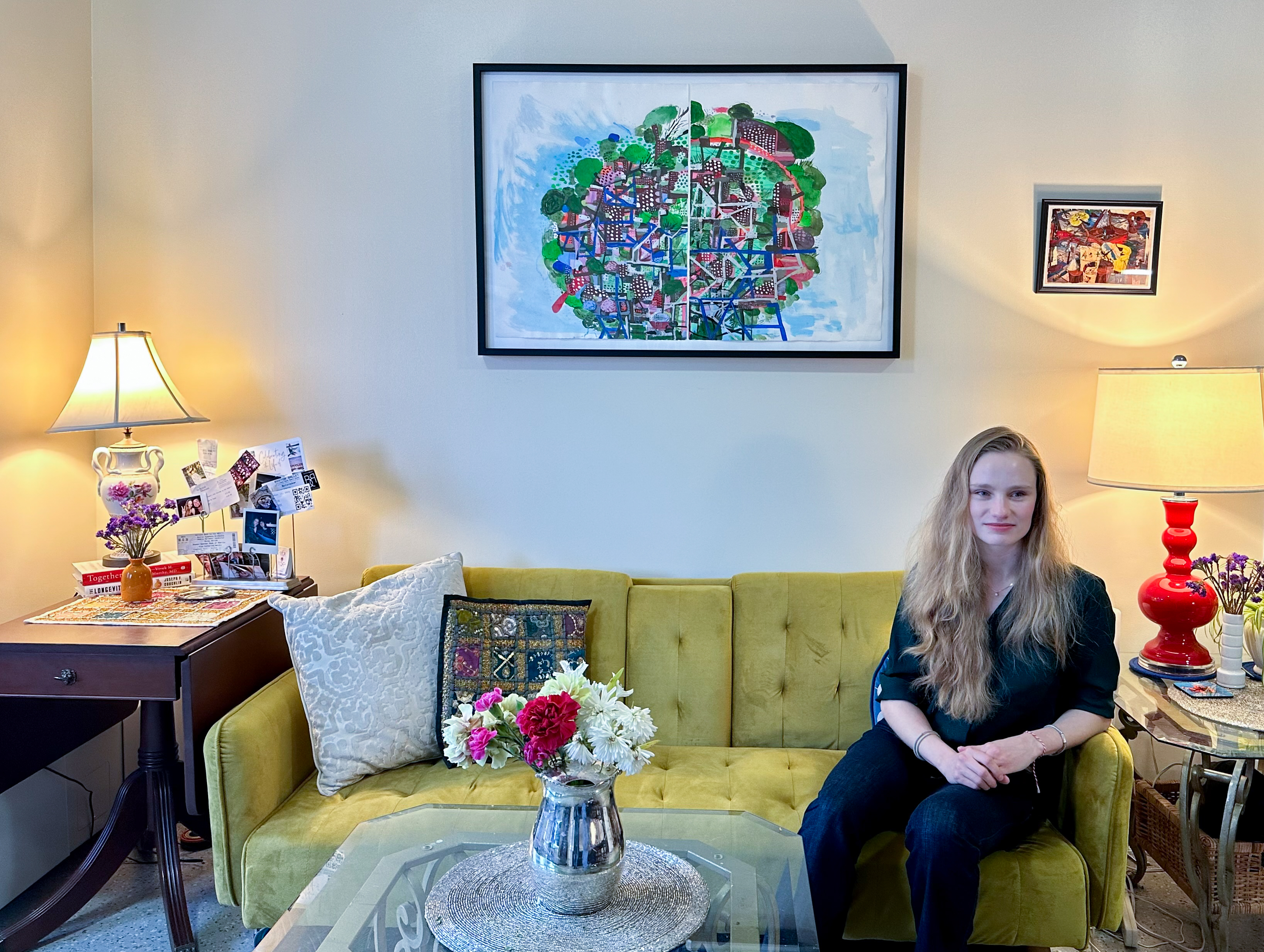 A woman sits on the right side of a green couch behind a coffee table with a vase of red and white flowers and underneath a black framed painting hanging on the wall. There is a lamp on end tables at either side of the couch.