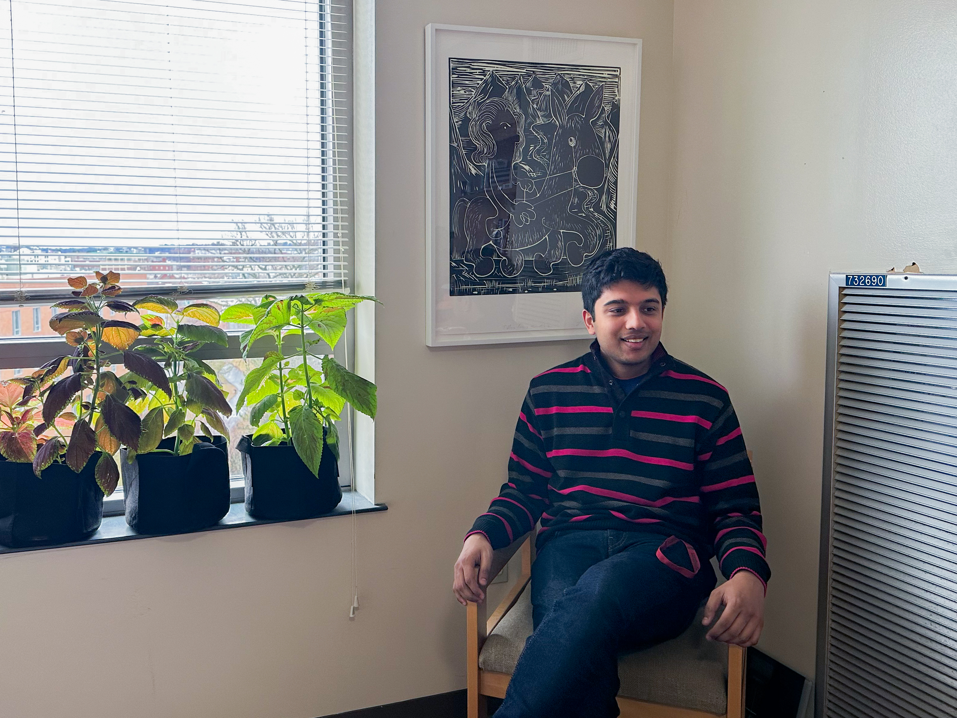 Man sitting in a chair in front of a large framed artwork mounted onto a wall to the left of a window with plants on the windowsill.