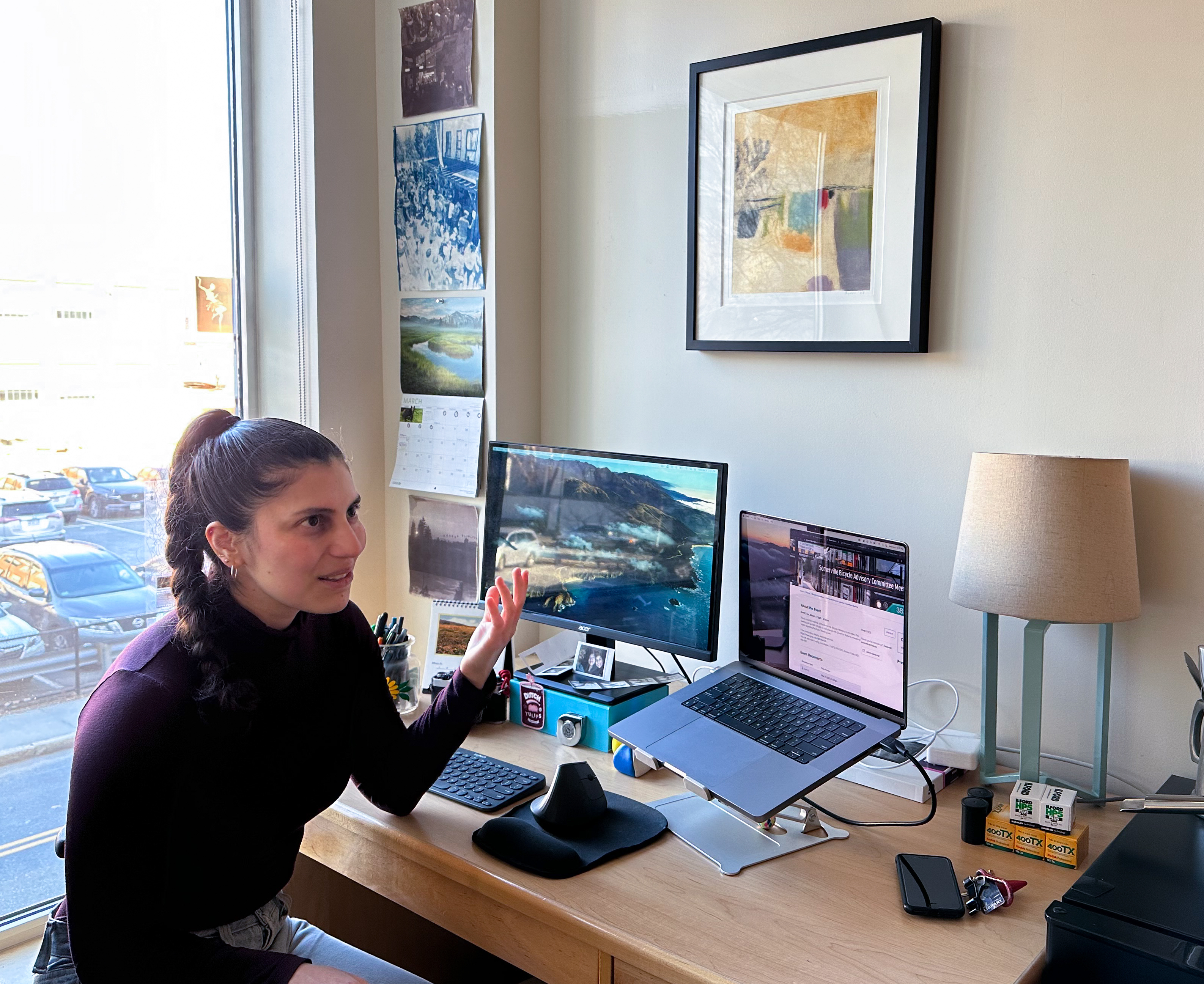 A woman in sitting next to a window in front of a wooden desk that holds two computers and a desk light. There is a black framed artwork hanging on the wall above the desk.