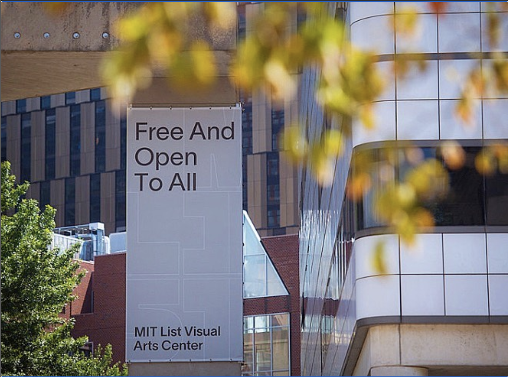 A large rectangular gray sign is visible through blurry leaves on a tree branch in the foreground. The text on the sign reads &quot;Free And Open To All MIT List Visual Arts Center&quot;