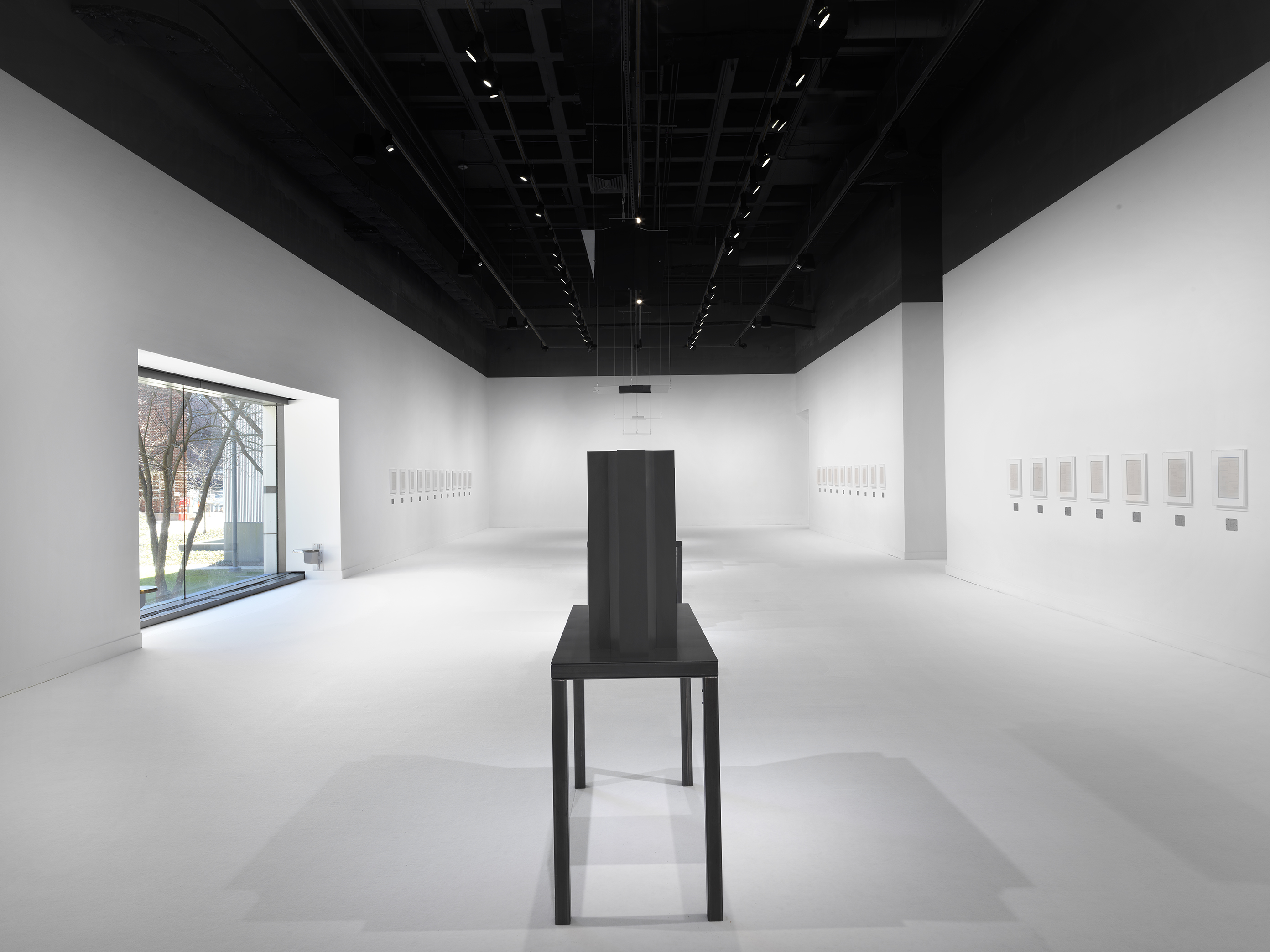 Flat plaster artworks in white frames are hung horizontally across a white-walled and floored gallery with a dark steel table and geometric sculpture in the center.