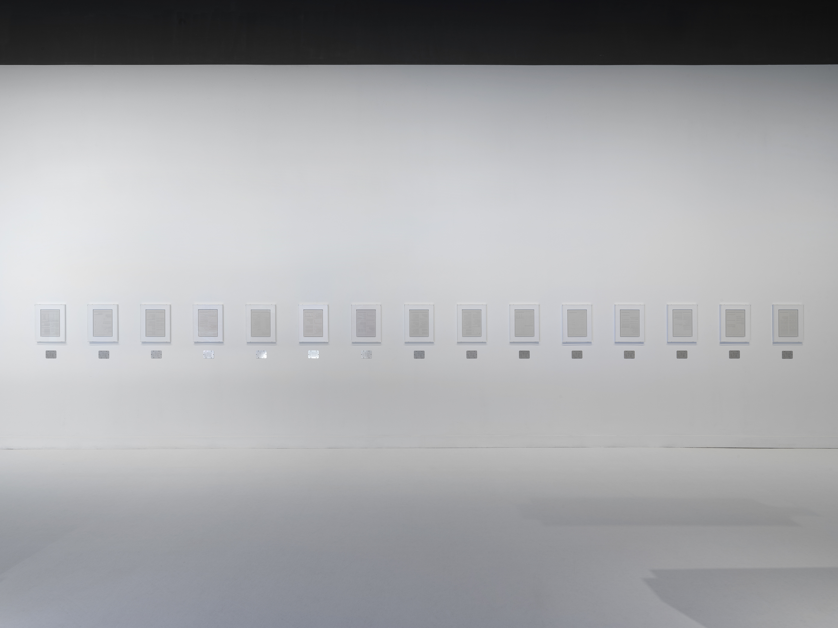 Fifteen page-sized plaster works in white frames are hung horizontally across white gallery walls, each with a small steel plaque below it.