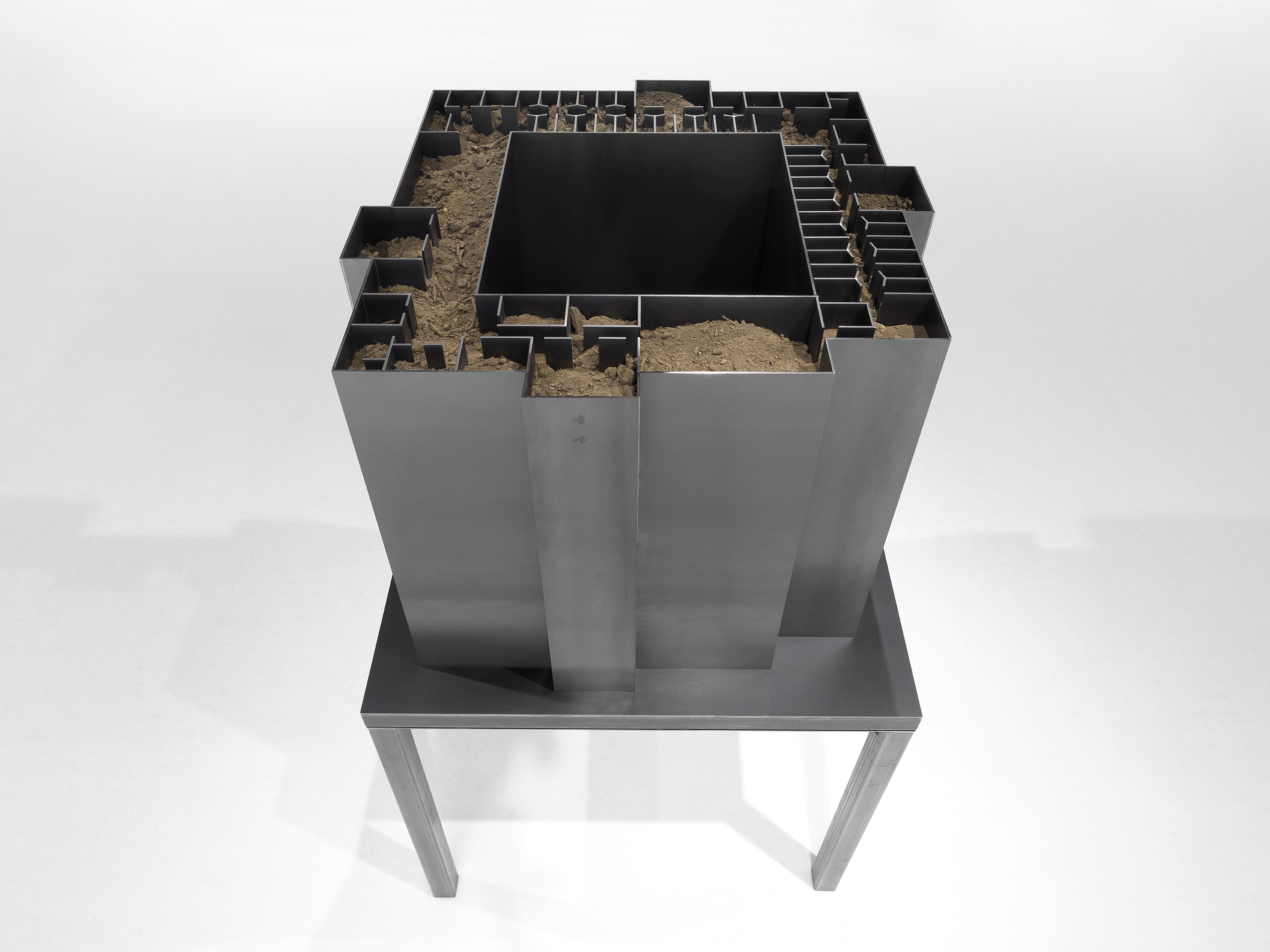 An aerial view of a dark, hollow steel sculpture with geometric inner walls is filled with dry dirt.