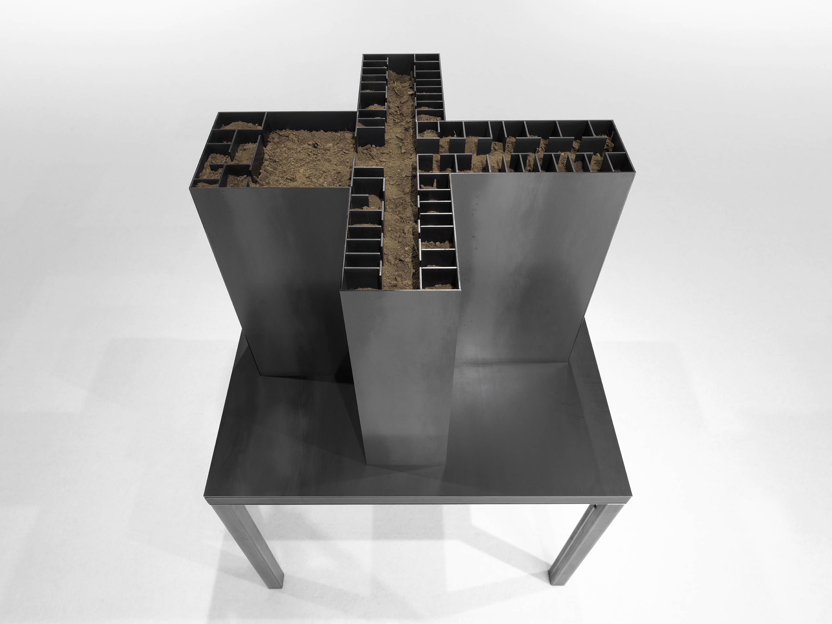 An aerial view of a dark, hollow steel sculpture with geometric inner walls is filled with dry dirt.