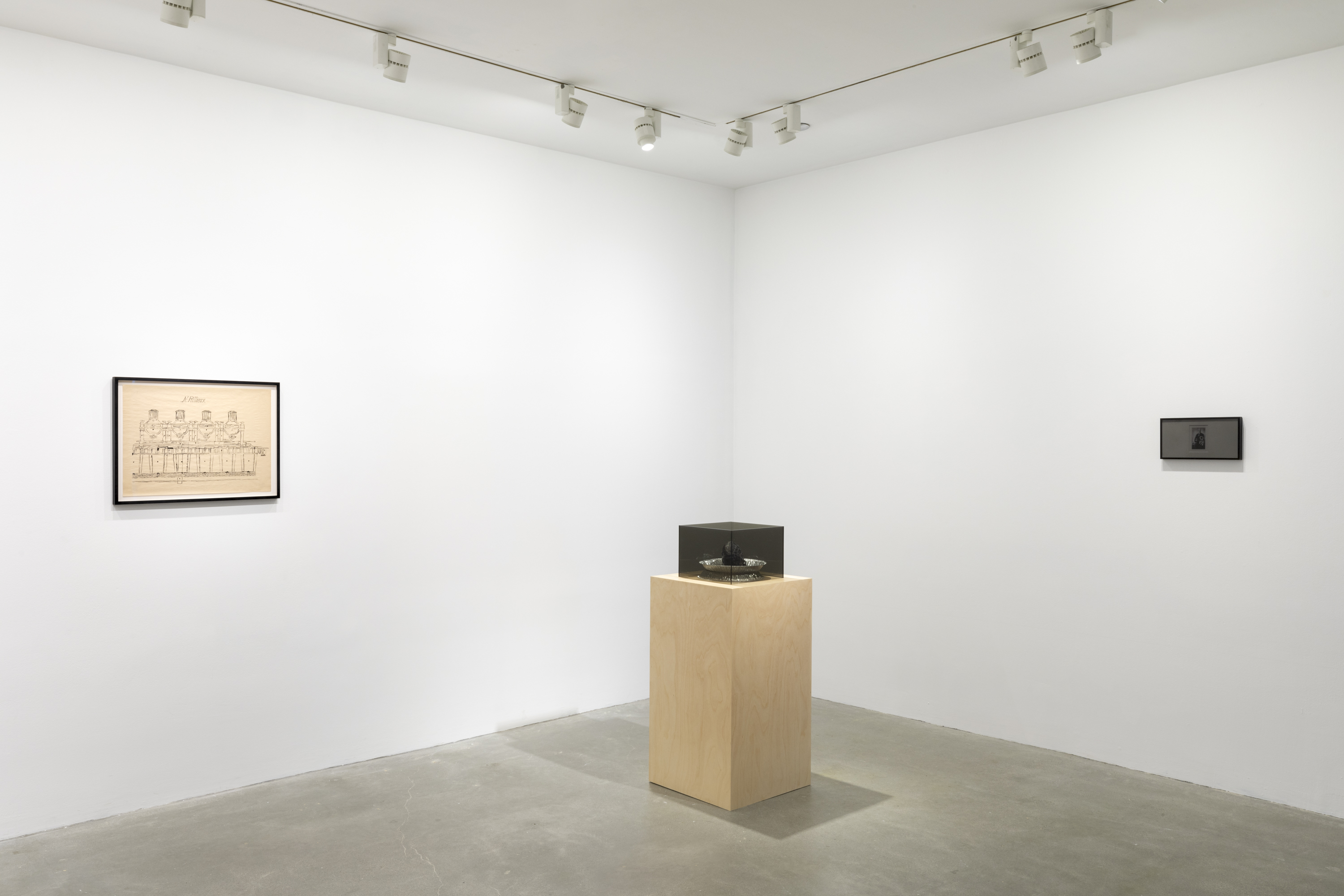 A white walled room with a wooden pedestal with a gray Plexiglas cube on top in the middle. To the left of the pedestal there is a black framed drawing hung on the wall. To the right of the pedestal there is a black and white printed image of Norbert Rilliuex behind a privacy filter with black framing.
