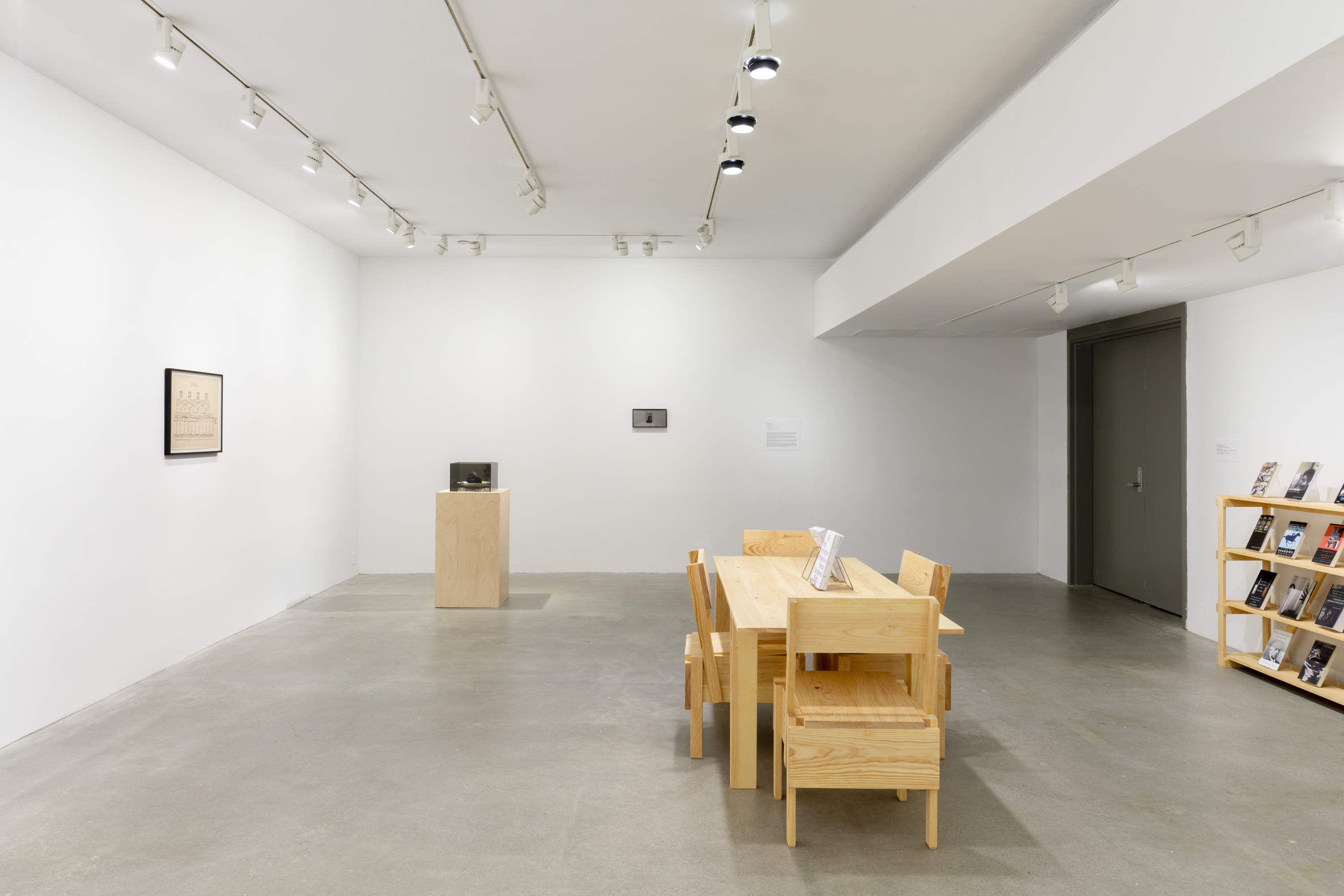 A white walled room with a tan wooden table is visible with four wooden chairs in the center. A wooden pedestal with a gray Plexiglas cube on top is visible to the left of the table in the middle of the room. To the right of the pedestal there is a black and white printed image of Norbert Rilliuex behind a privacy filter with black framing. To the left of the pedestal there is a black framed drawing hung on the wall. A tan wooden bookshelf is partially visible on the right side of the image.
