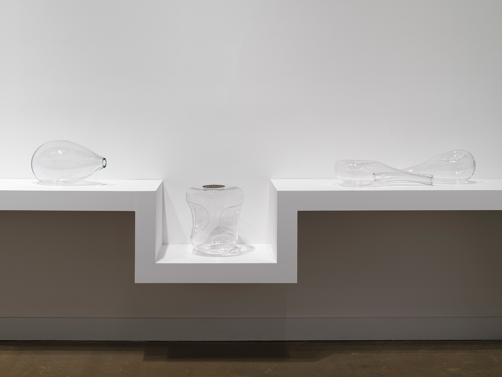 Four glass sculptures sit on a shelf against a white wall. They resemble stretched, deformed, or indented vessels. The shelf sinks to accommodate a bulkier sculpture, the top of which is level with the rest of the shelf. Its flat top contains a round drain cover, like one you would find in a shower.