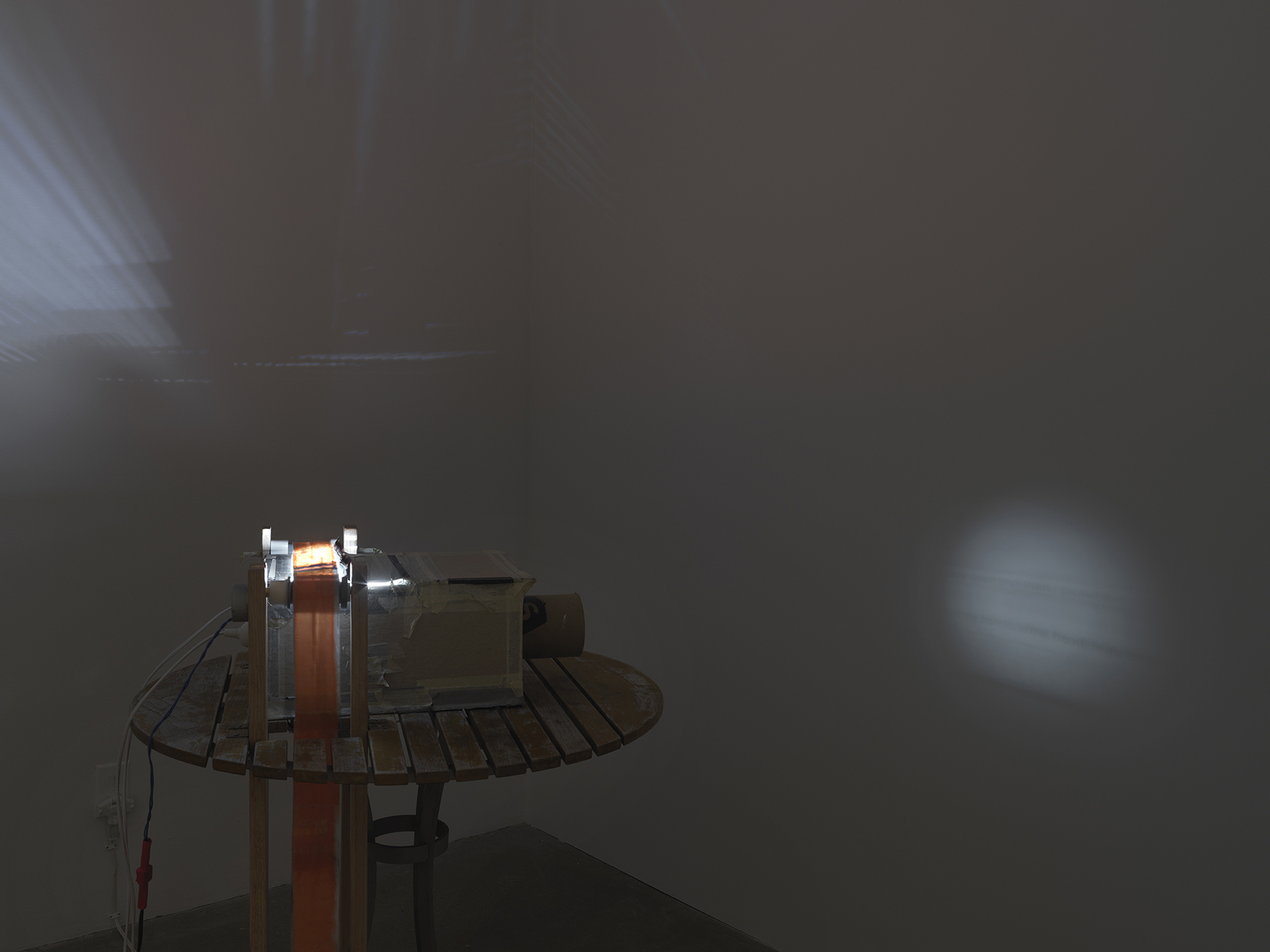 A boxy makeshift projector sits on a wooden table; it is crudely constructed with materials like cardboard and duct tape. Three cords are attached to its back, and a strip of semi-transparent material runs through the device, like a film strip would. A circle of light with a blurry strip of text is projected through the projector’s cylindrical “lens.” A more abstract pattern of light rays is also thrown across an adjacent wall.