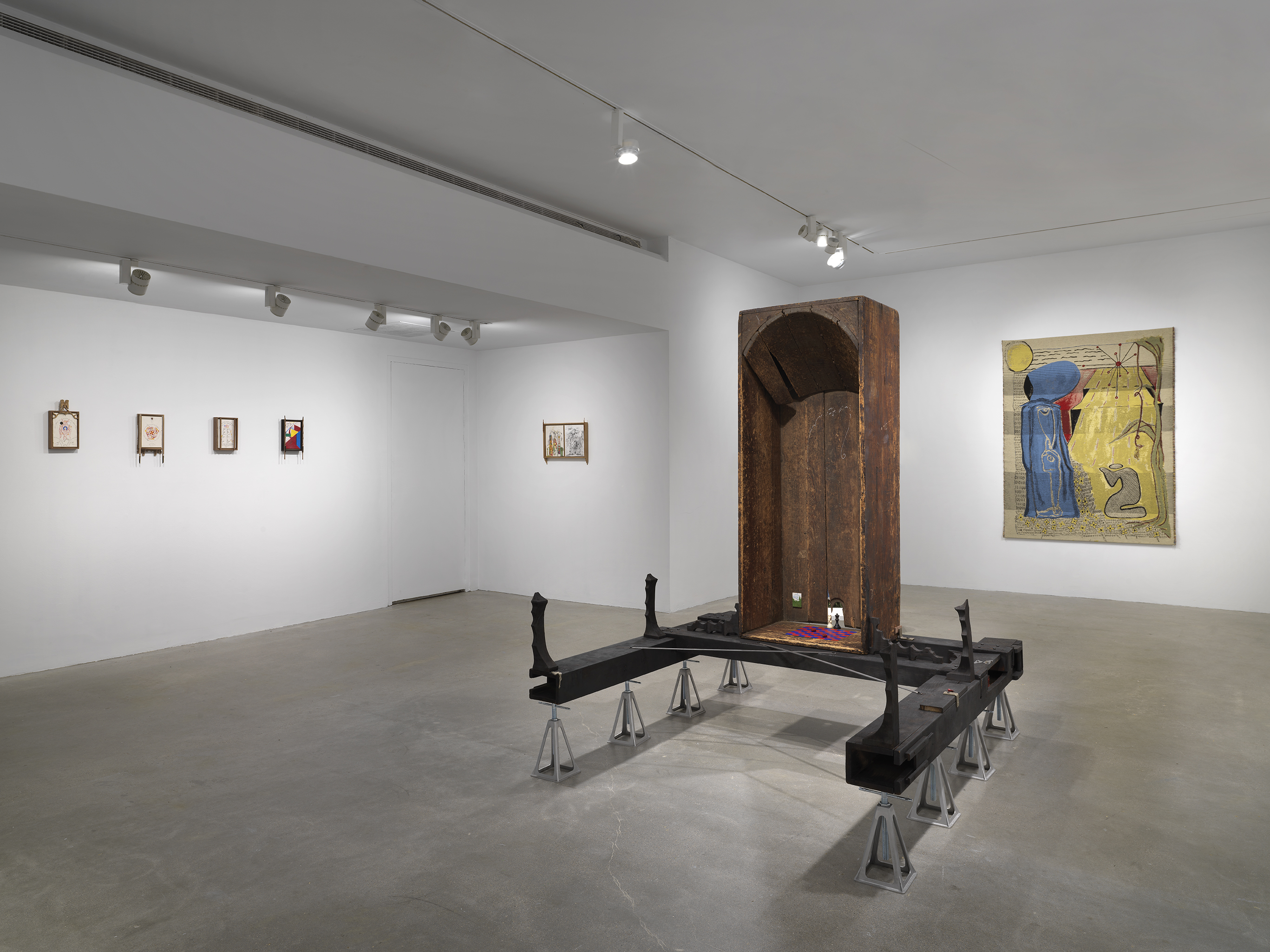In the foreground, a vertical wooden bathtub sits on horizontal wooden beams, which are held up by eight metal supports. Inside the bathtub are small painted scenes and a red and blue checkerboard pattern on the base. On the left walls, five small drawings hang in wooden frames. In the rear, a golden tapestry depicts two figures, one standing and one kneeling.