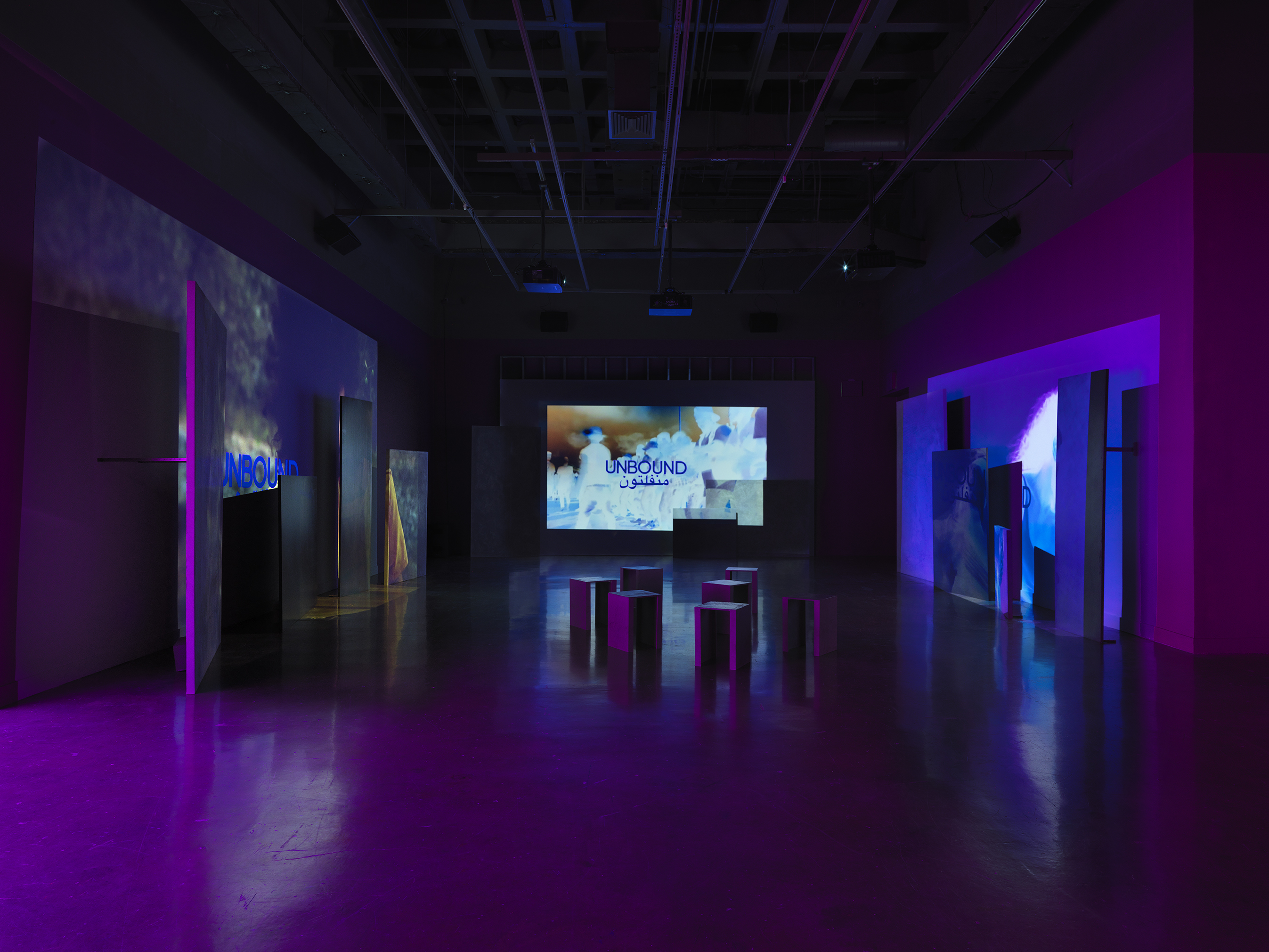 Videos are projected onto uneven surfaces of large panels against three walls. The central projection shows glowing, almost psychedelic inverted footage of a group of figures in a line. The right projection shows a woman (also in unnatural, inverted tones) dancing, and the left projection shows a person wearing a yellow shirt. All three images are overlaid with text in English and Arabic reading, “Unbound.”
