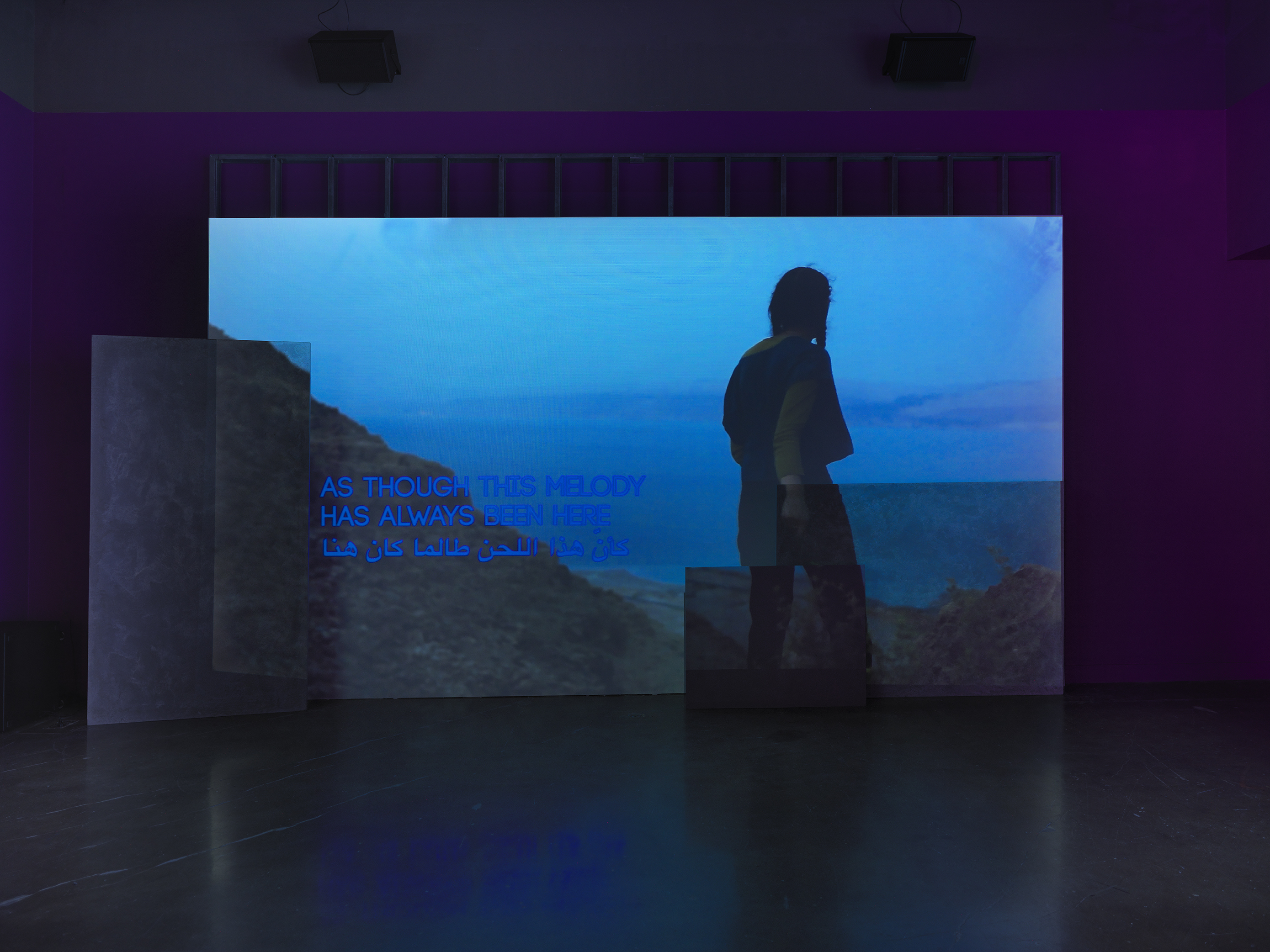 A deep blue video depicting a person looking out from a mountain onto a body of water is projected onto an uneven surface of overlapping rectangular panels. Overlaid on the video footage, English and Arabic text reads “As though this melody has always been here”.